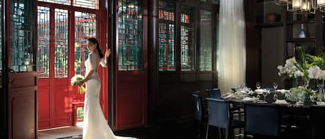 A bride in a white dress standing in a room with traditional Chinese doors and a dining area set up for a wedding reception.