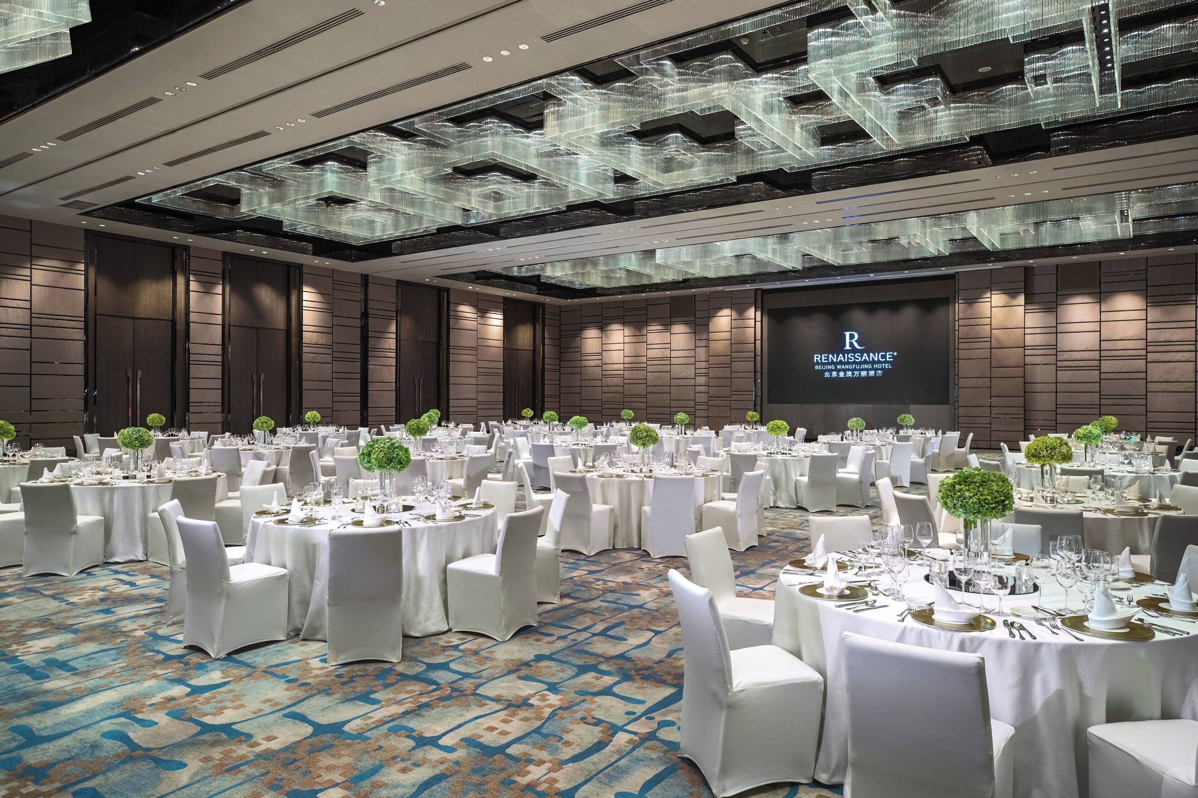 A banquet hall with round tables set for a formal event, featuring a modern ceiling design and a large screen displaying the hotel name.