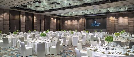 A banquet hall with round tables set for a formal event, featuring a modern ceiling design and a large screen displaying the hotel name.