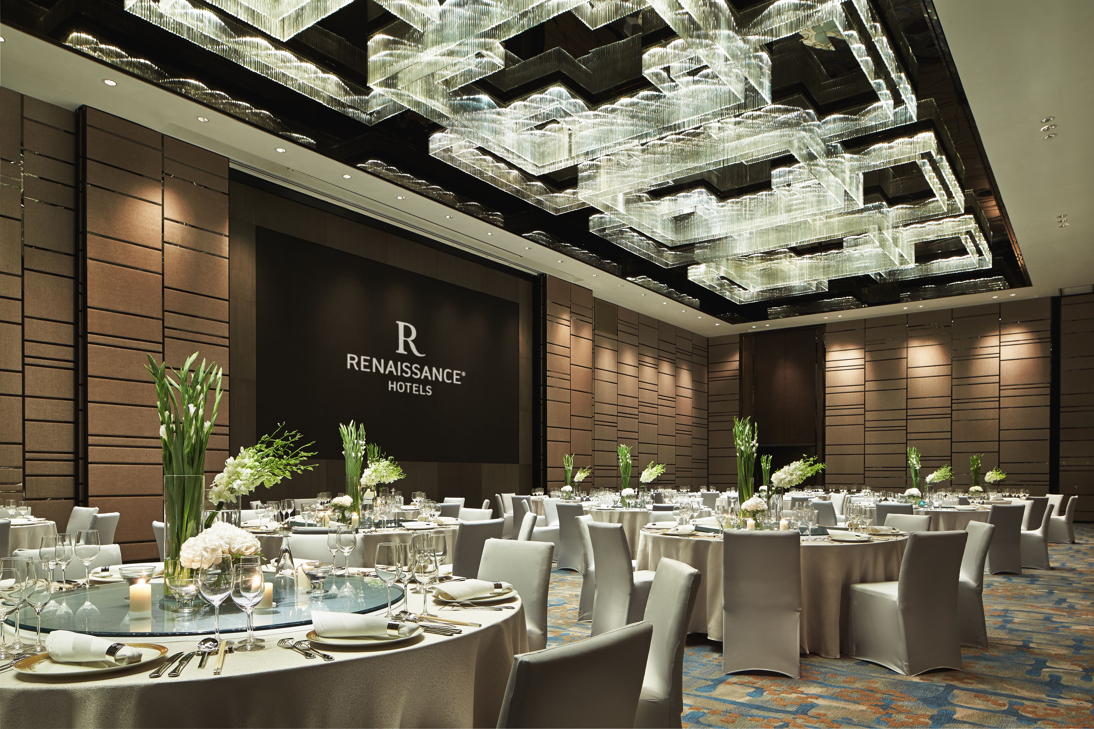 A banquet hall with round tables set for dining, a large screen displaying the hotel name, and a modern chandelier.