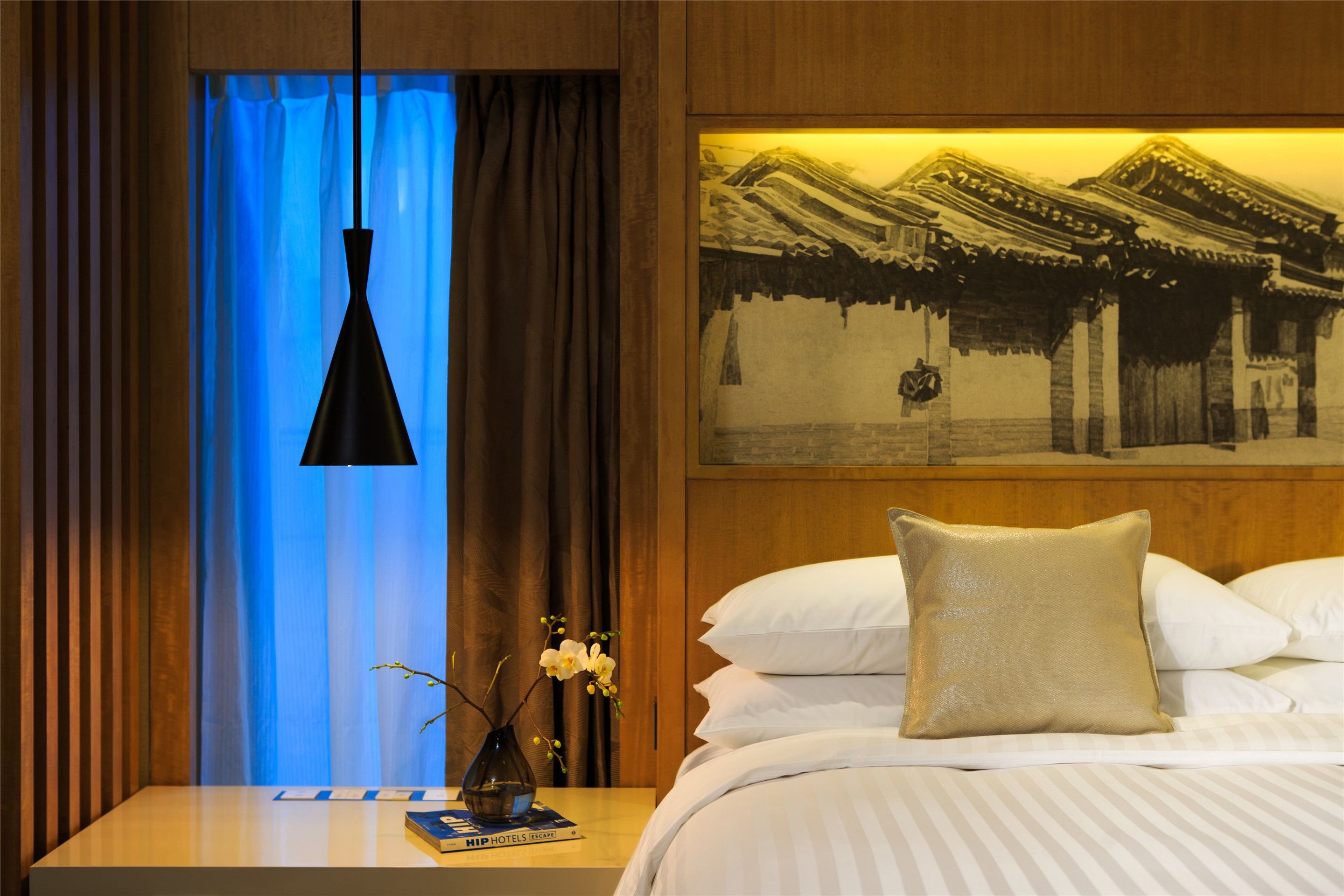 Ein Hotelzimmer mit Bett, Nachttisch, einer Blumenvase, einem Bild an der Wand und einem blau beleuchteten Fenster.