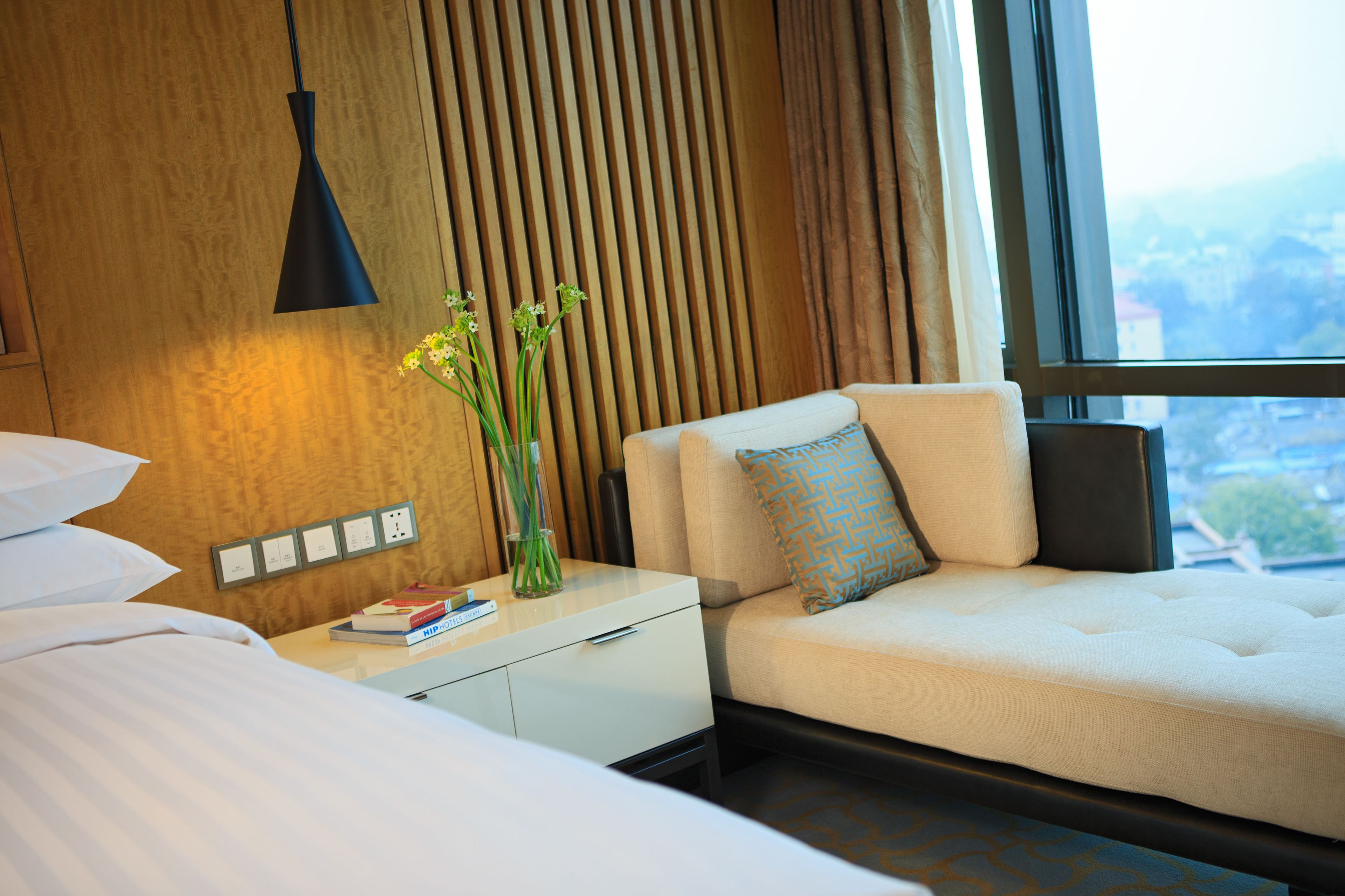 A modern hotel room with a bed, a sofa, a nightstand with a lamp, and a window offering a city view.