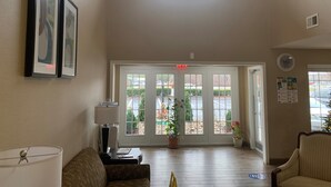 A spacious lobby with a high ceiling, a chandelier, framed artwork, a lamp, a sofa, and a view through large glass doors to an outdoor area.