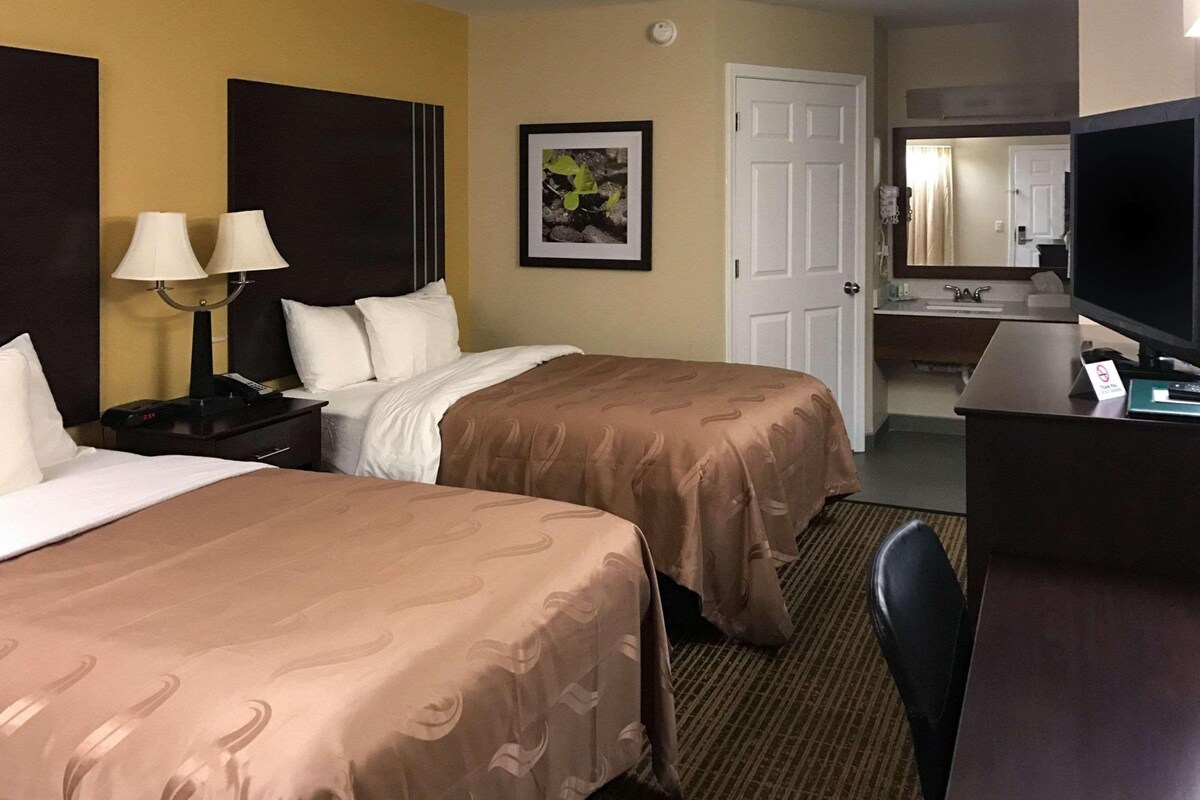A hotel room with two beds, a desk with a television, a bathroom with a mirror, and a framed picture on the wall.
