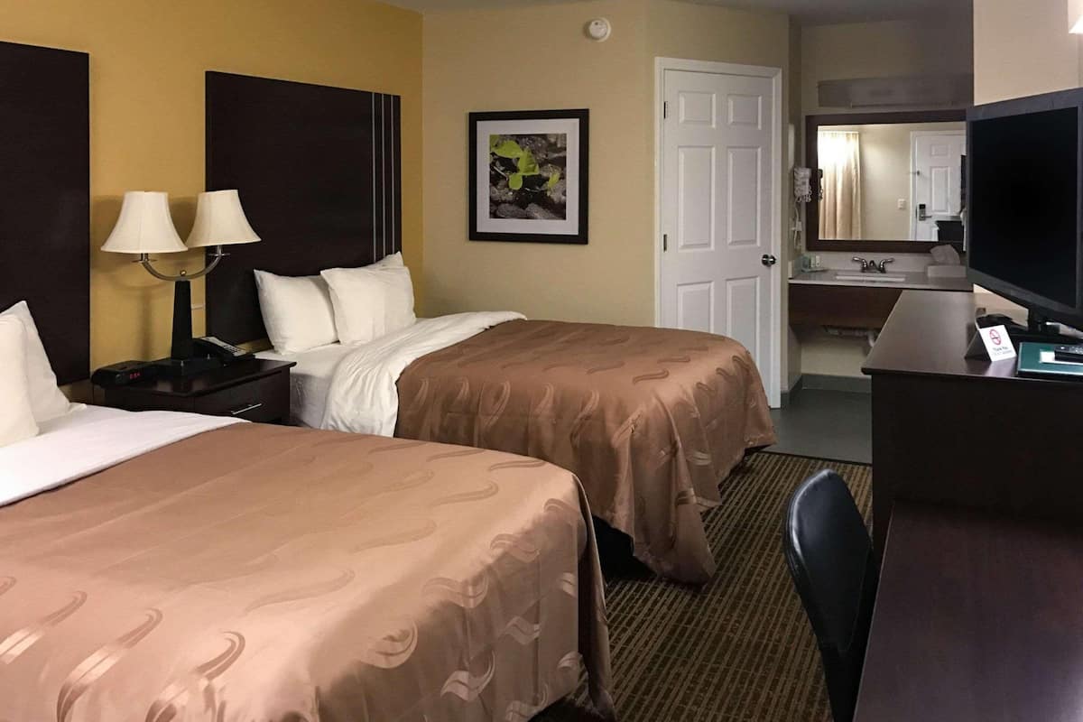 A hotel room with two beds, a desk with a television, a bathroom with a mirror, and a framed picture on the wall.