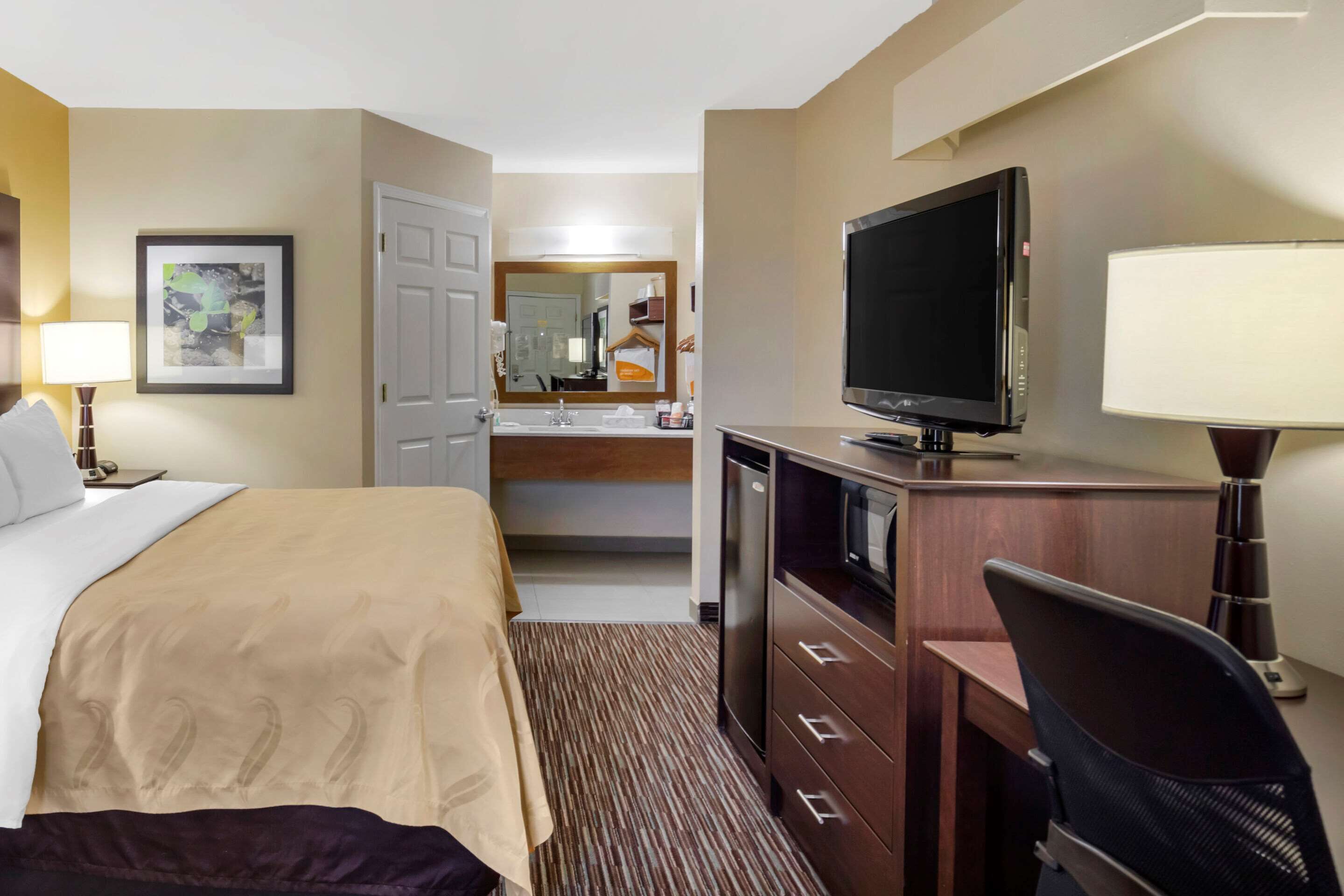 A hotel room with a bed, a desk with a computer, a TV, a mirror, and a bathroom visible in the background.