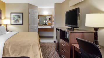 A hotel room with a bed, a desk with a computer, a TV, a mirror, and a bathroom visible in the background.
