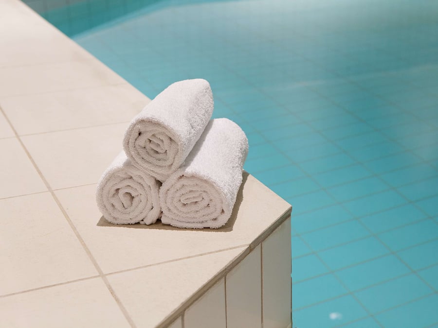 Three rolled white towels stacked on a white tiled ledge by a pool.