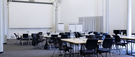 Une salle de conférence dotée d’un grand écran blanc, de plusieurs tables avec des chaises et d’un tapis à motifs.