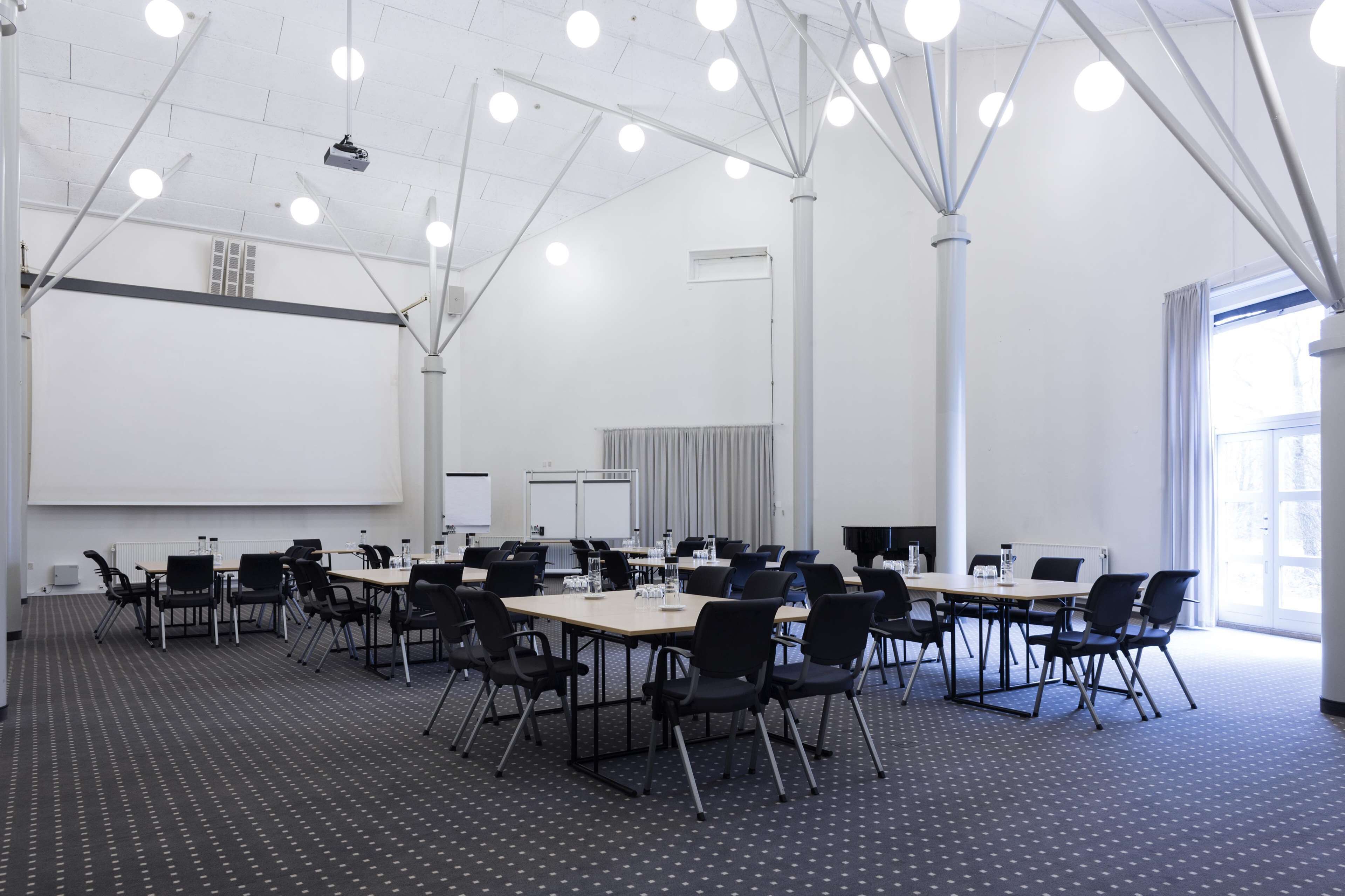 A conference room with multiple tables and chairs, a projector screen, and a large window.