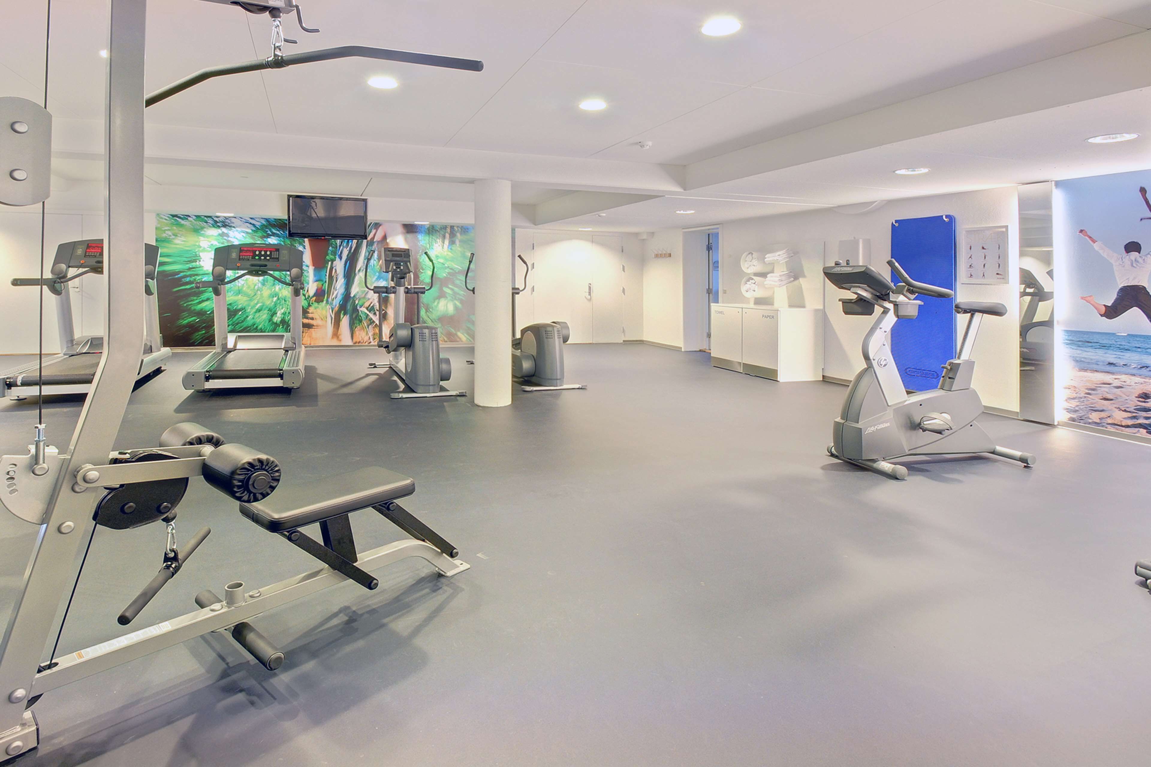 A modern gym with cardio machines, weight training equipment, and a large screen displaying exercise videos.