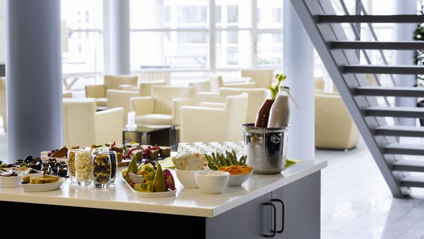 A buffet table with various food items and a drink in a bucket, set in a bright room with large windows and a staircase. - Scandic Sønderborg (Sønderborg)