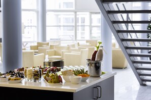 A buffet table with various food items and a drink in a bucket, set in a bright room with large windows and a staircase. - Scandic Sønderborg (Sønderborg)