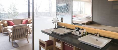 A modern bathroom with two sinks, a large mirror, and a seating area visible through a glass partition.