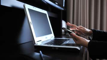A laptop on a desk with a person holding a smartphone.