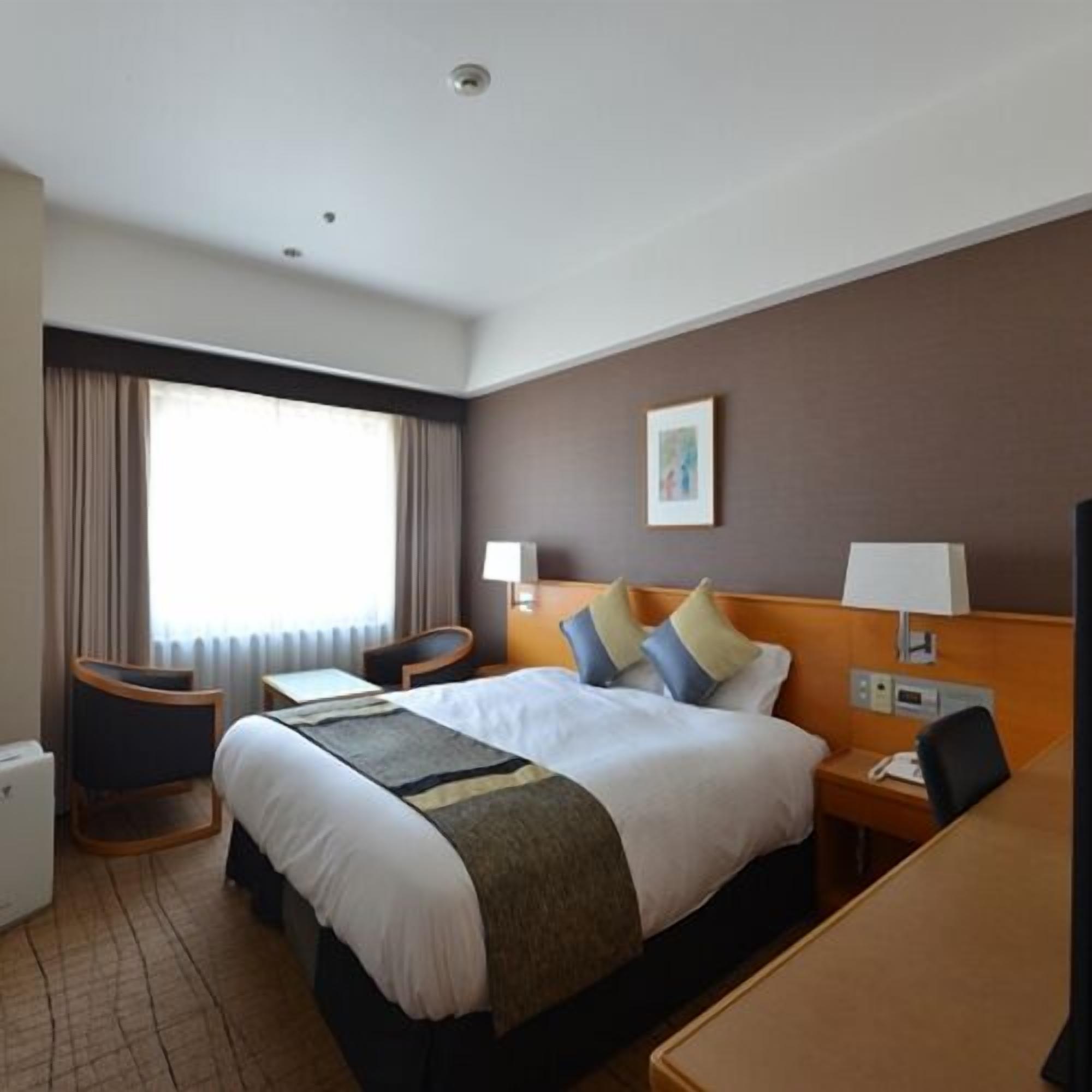 Una habitación de hotel con una cama, mesitas de noche, un escritorio, una silla, una ventana con cortinas y un cuadro colgado en la pared.