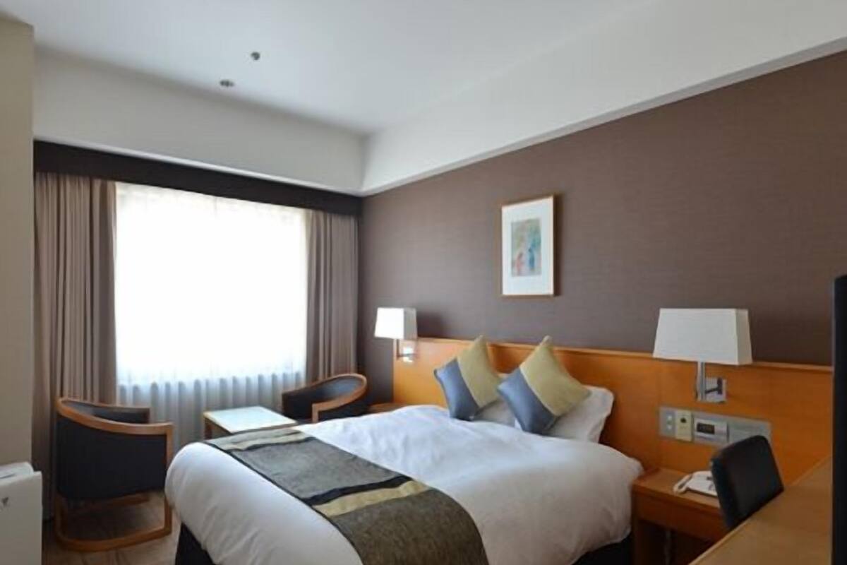 A hotel room with a bed, bedside tables, a desk, a chair, a window with curtains, and a wall-mounted picture.
