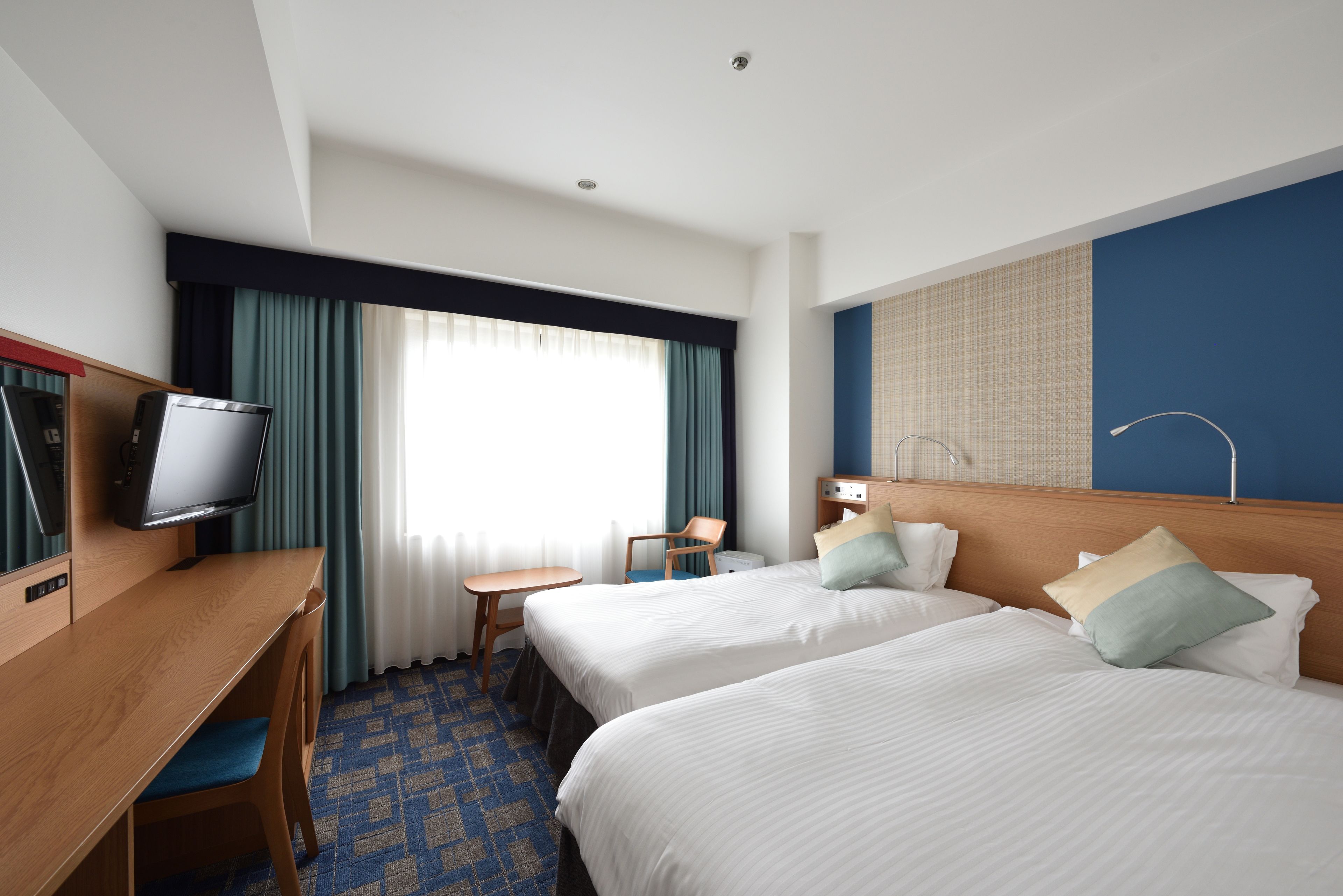 A hotel room with two beds, a desk with a television, a small table, and a window with curtains.