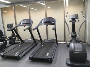 Three exercise machines in a gym with a grey carpet and a glass wall in the background. - Hotel Granvia Hiroshima (Hiroshima)