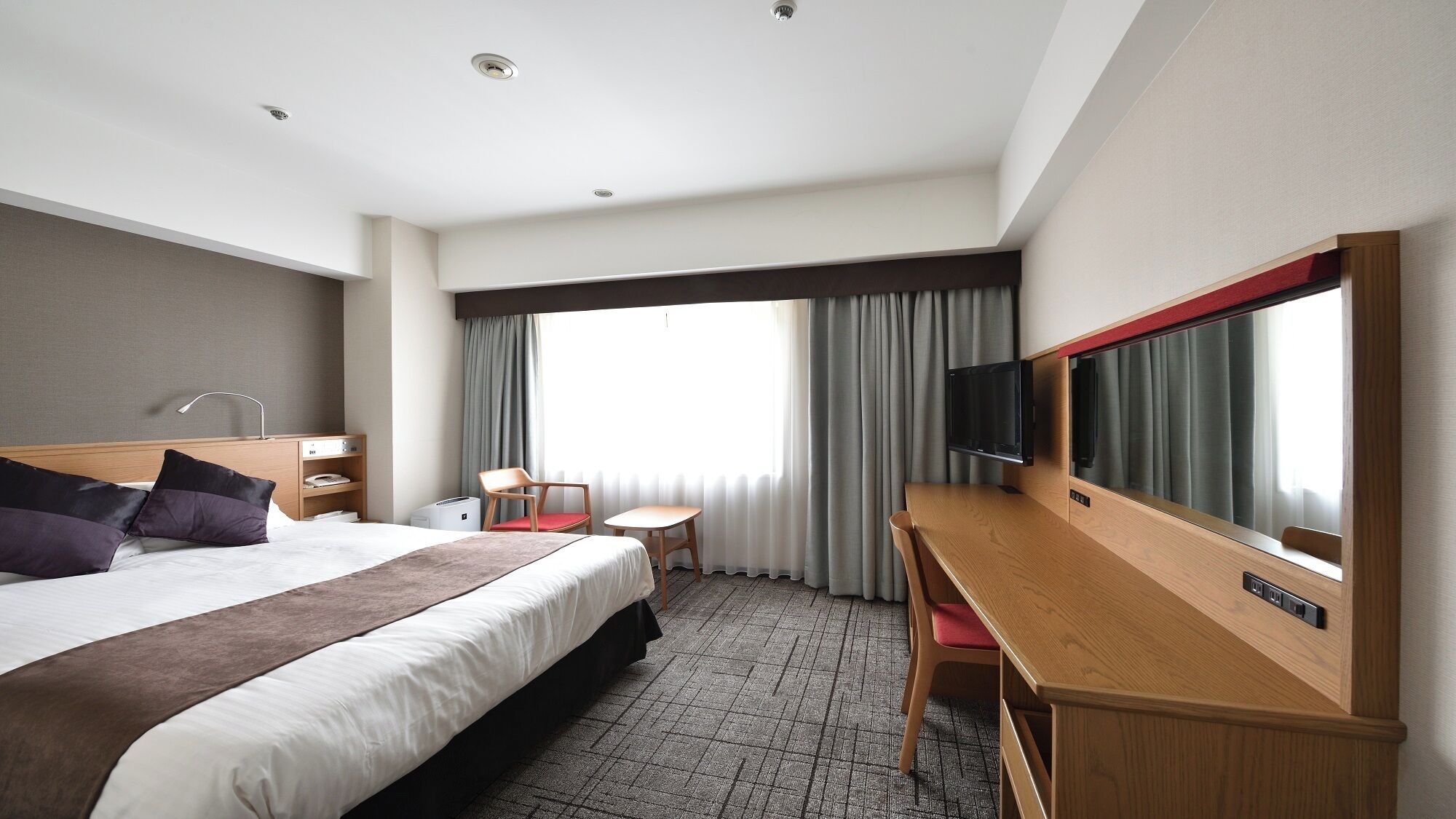 Habitación de hotel con cama, escritorio, silla, televisión y ventana con cortinas.