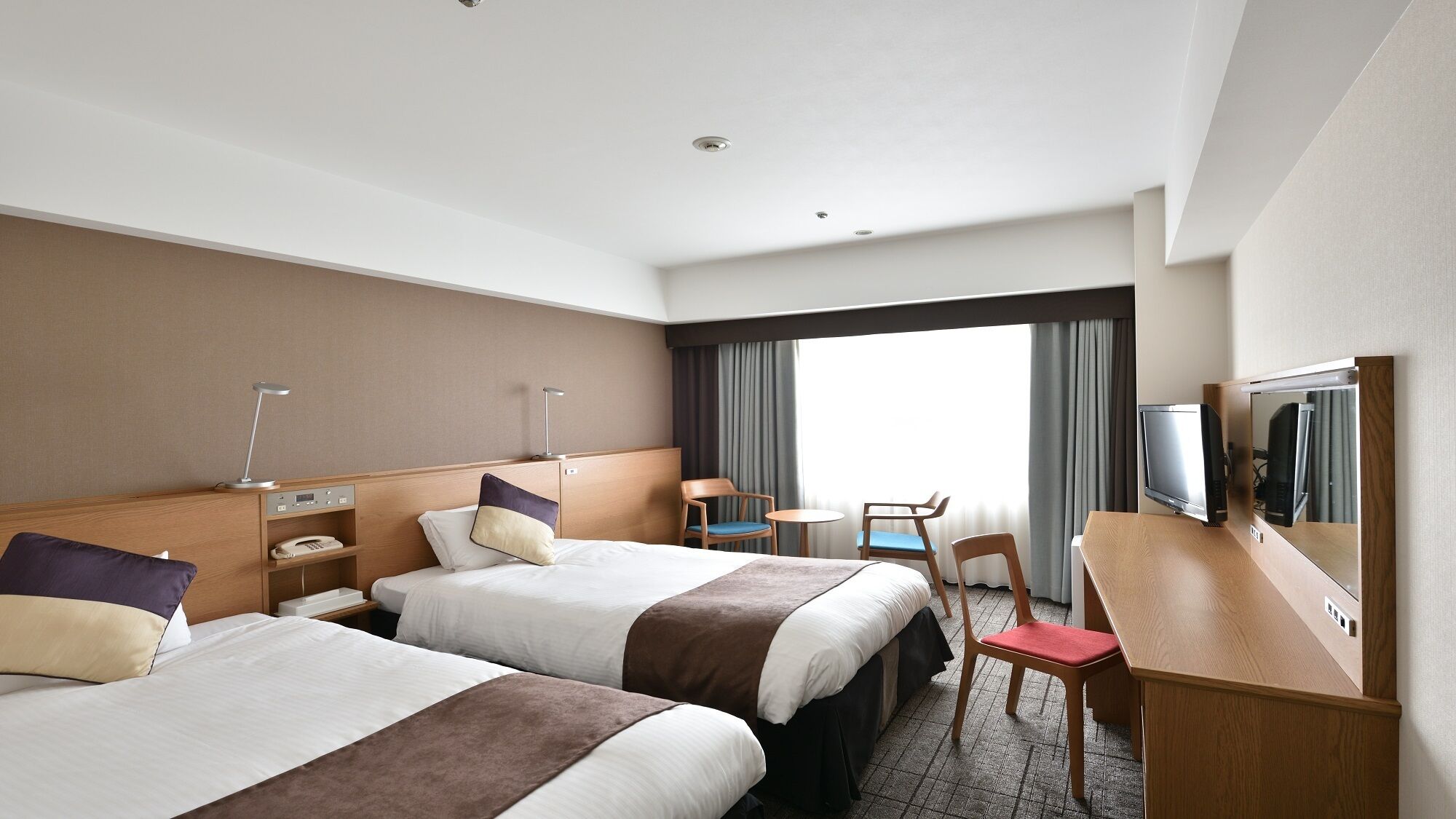 Une chambre d’hôtel avec deux lits, un bureau, une chaise, une télévision et une grande fenêtre.