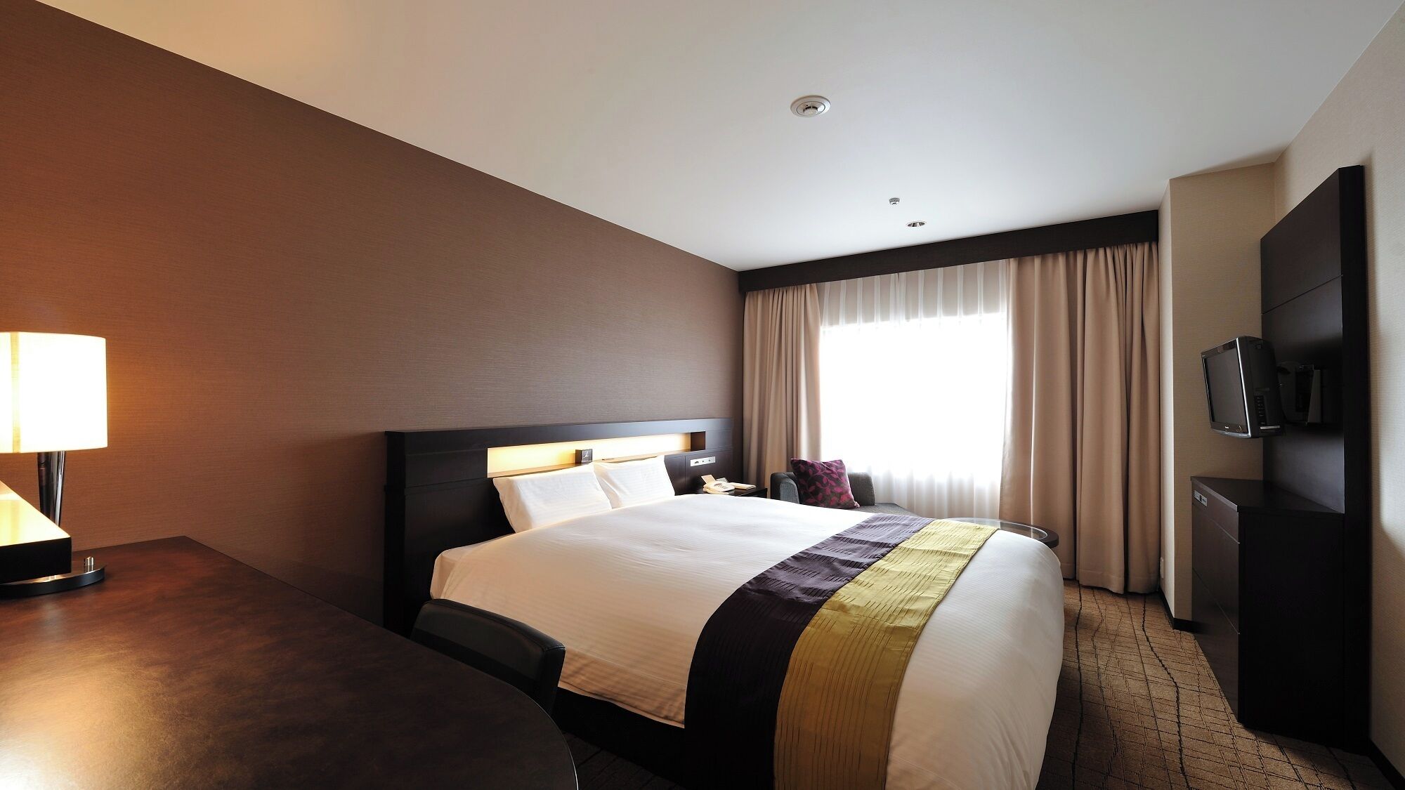 Una habitación de hotel con cama, escritorio, lámpara, televisor y ventana con cortinas.