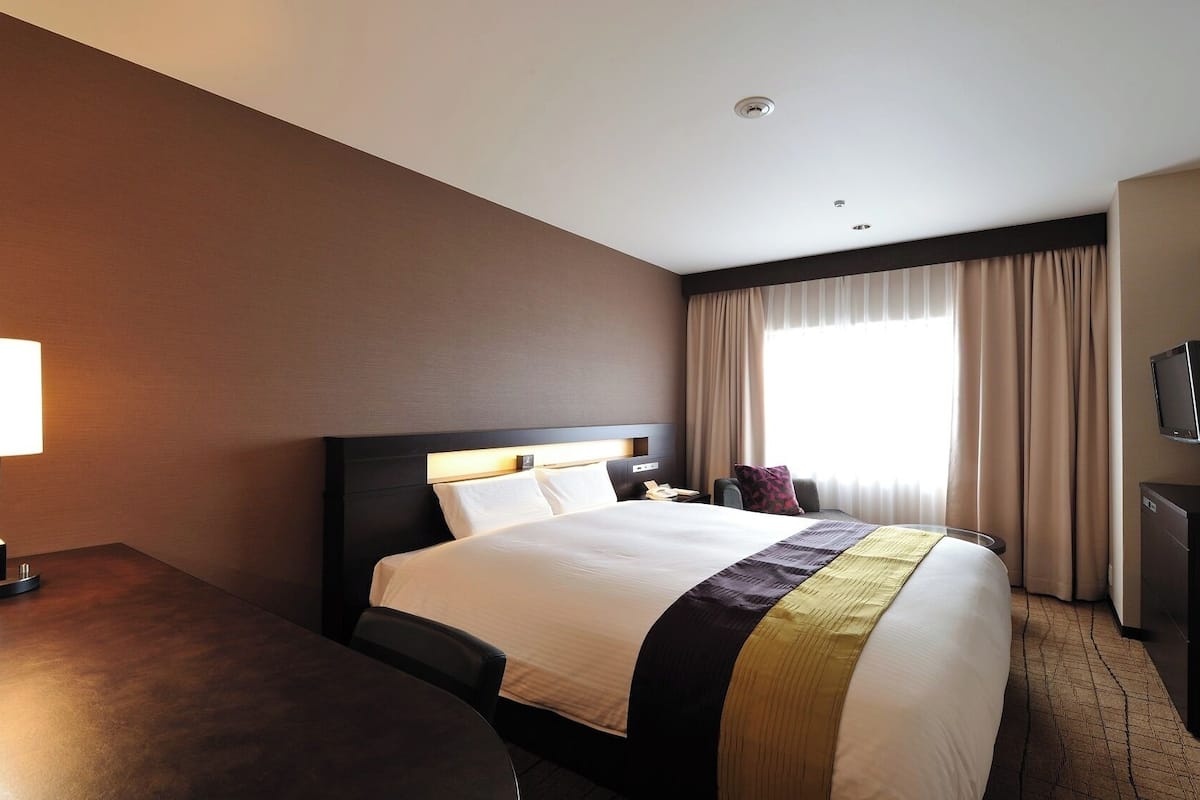 A hotel room with a bed, a desk, a lamp, a TV, and a window with curtains.