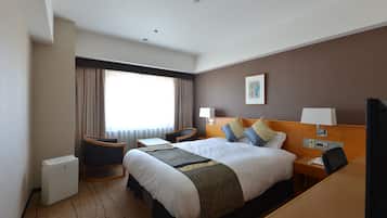 A hotel room with a large bed, a desk, a chair, a small table, a lamp, a picture on the wall, and a window with curtains.