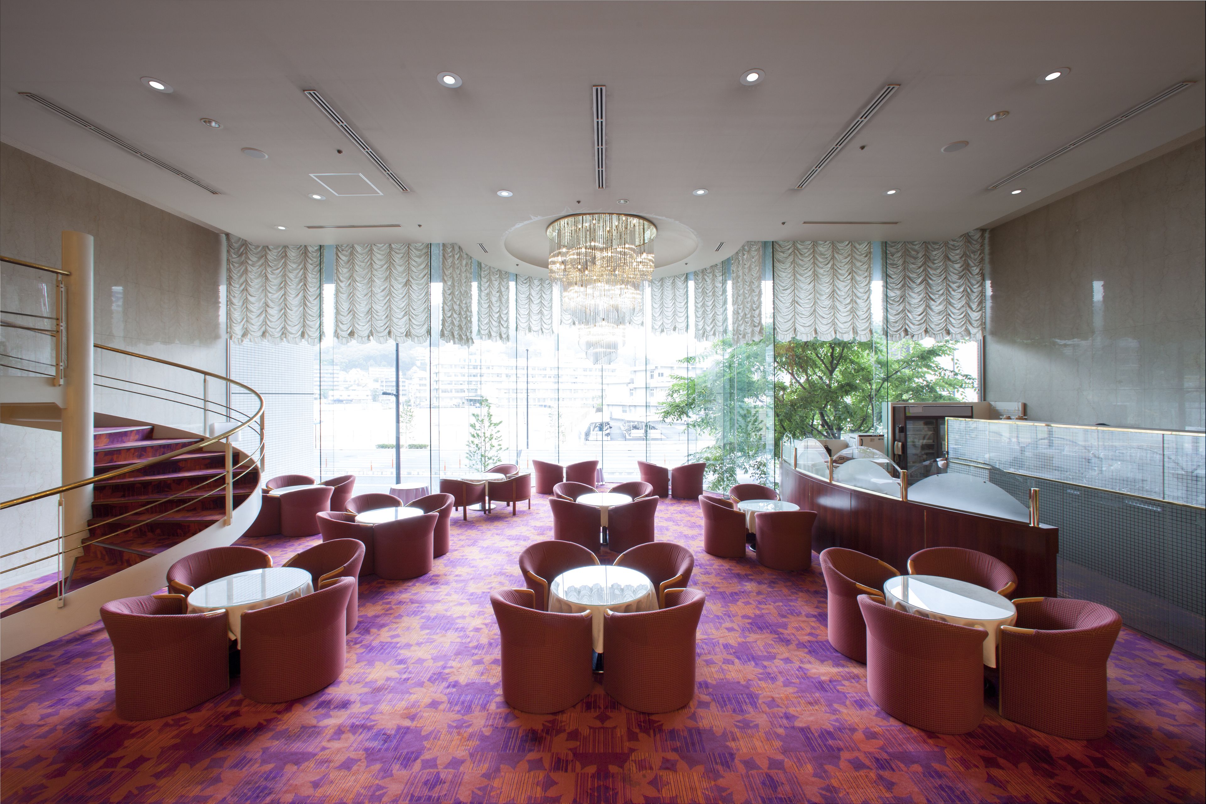 A hotel lobby with a central seating area, a staircase, and a view of the outdoors.