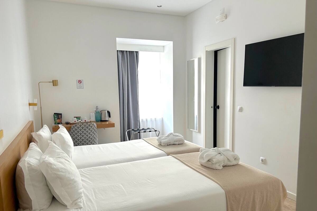 Habitación de hotel con dos camas, un televisor de pantalla plana, una ventana con cortinas, una mesita de noche con un hervidor y un perchero para toallas.