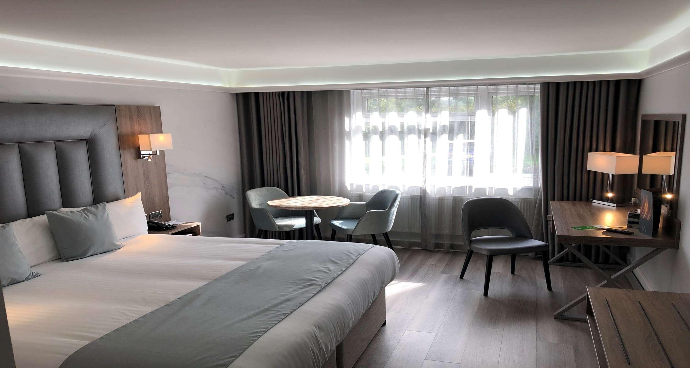 A modern hotel room with a large bed, a desk with a chair, a small round table, and a large window with curtains.