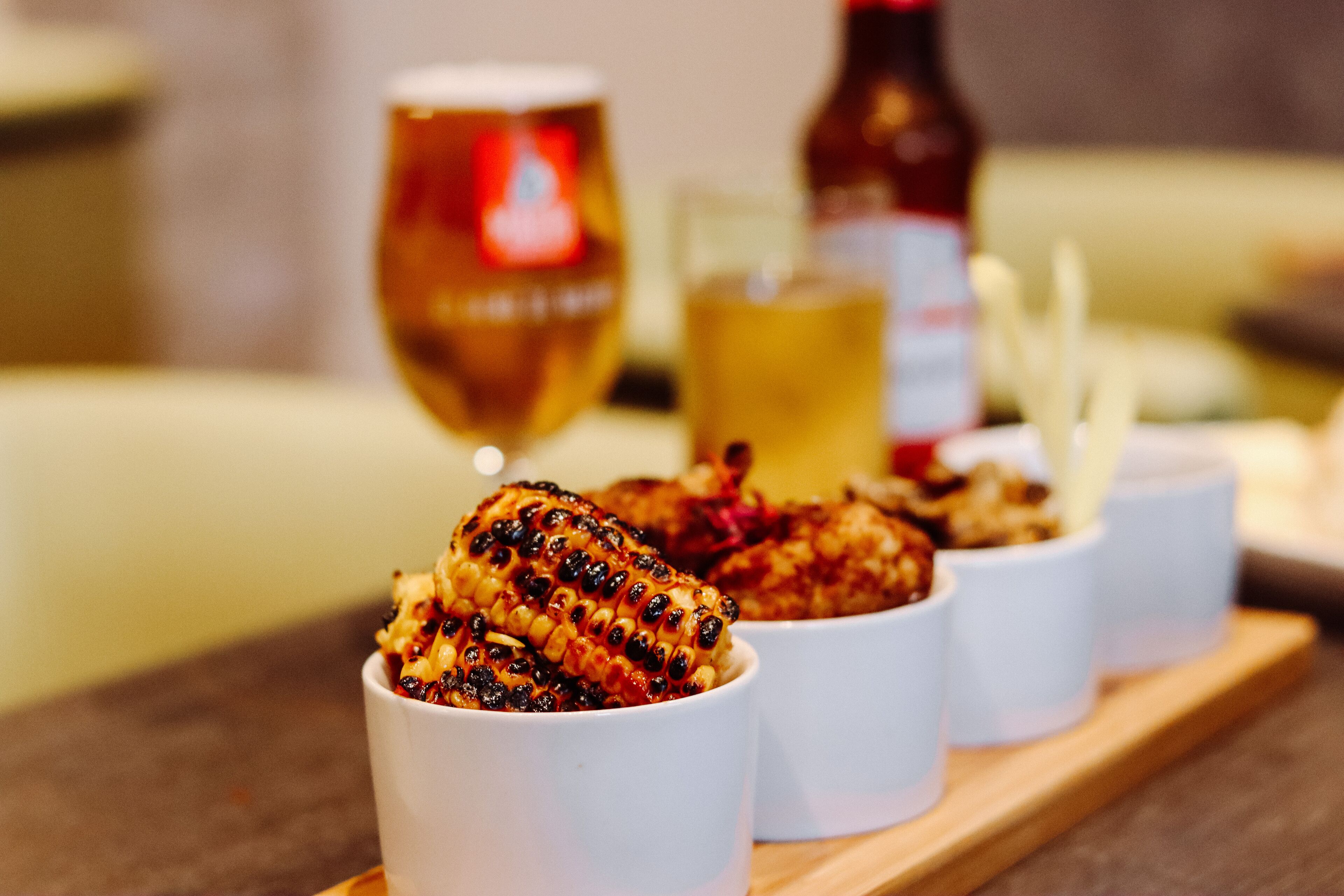 A wooden serving board with grilled corn on the cob and small white cups, likely containing sauces or dips, with a bottle and glass in the background.