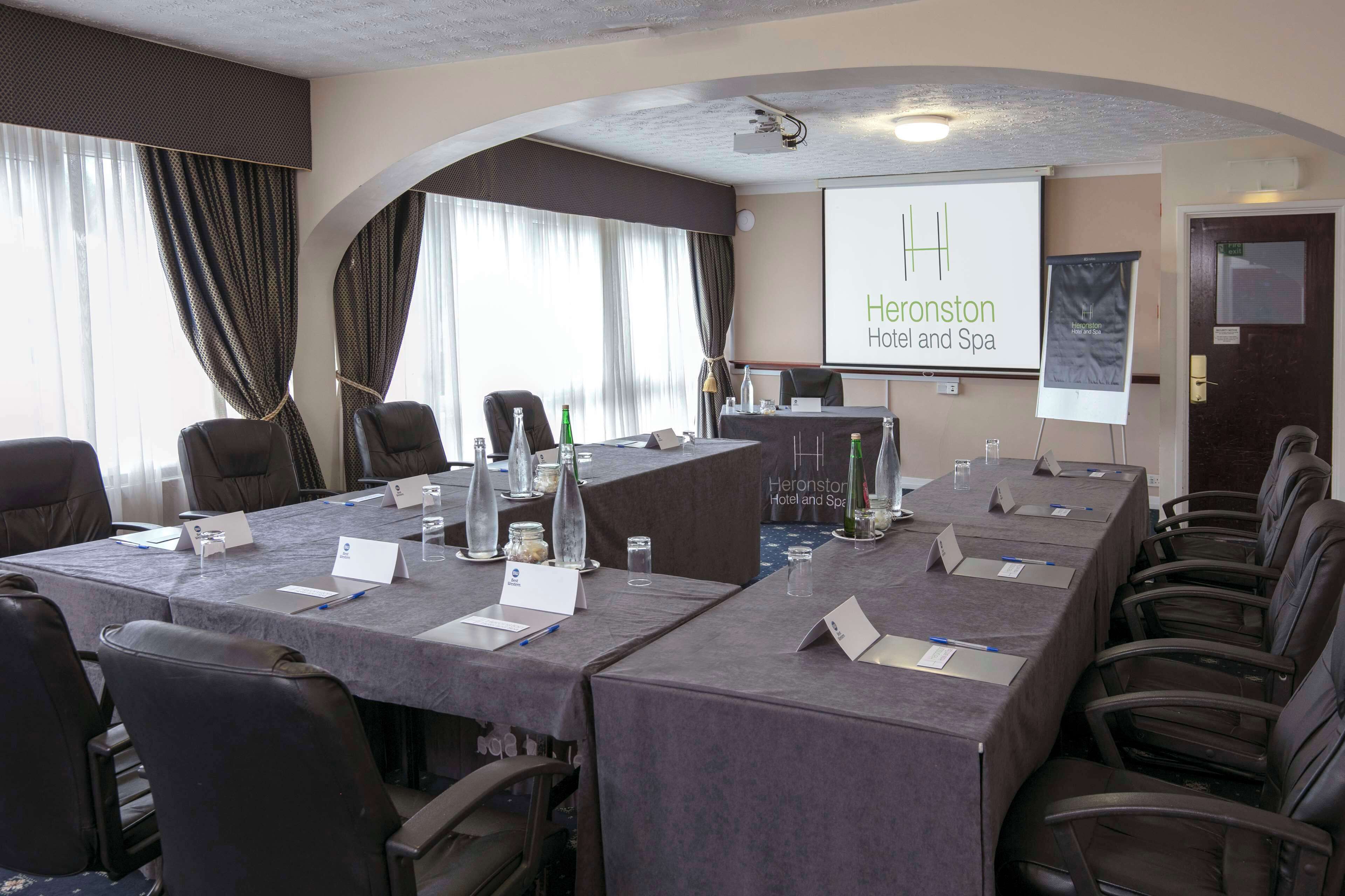 A conference room at Heronston Hotel and Spa with a long table, chairs, name cards, and a projector screen displaying the hotel name.
