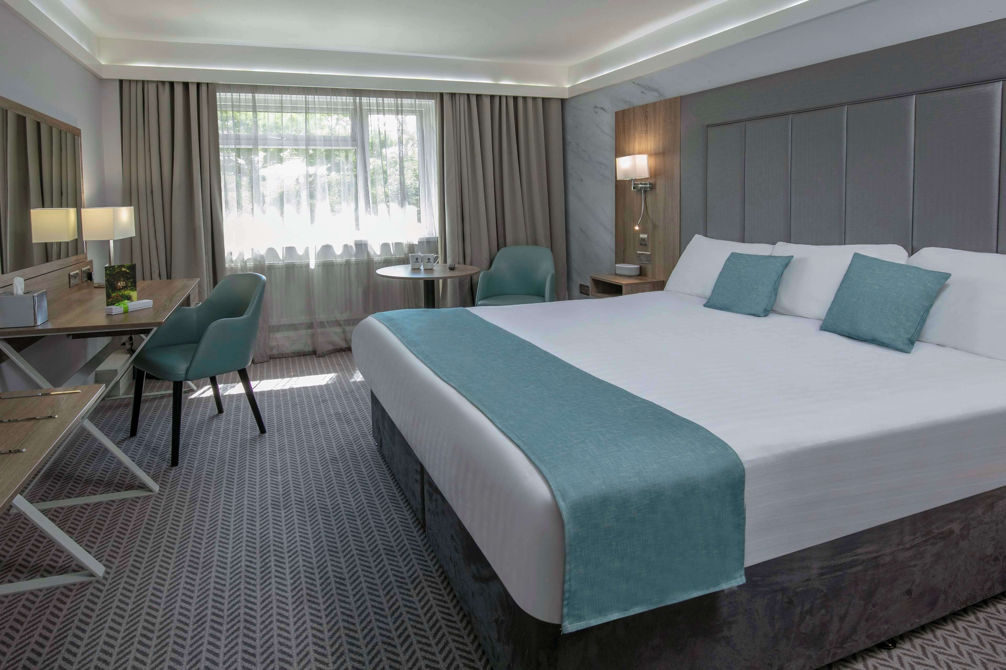 Habitación de hotel con una cama grande, un escritorio con silla, una mesita y un ventanal con cortinas.