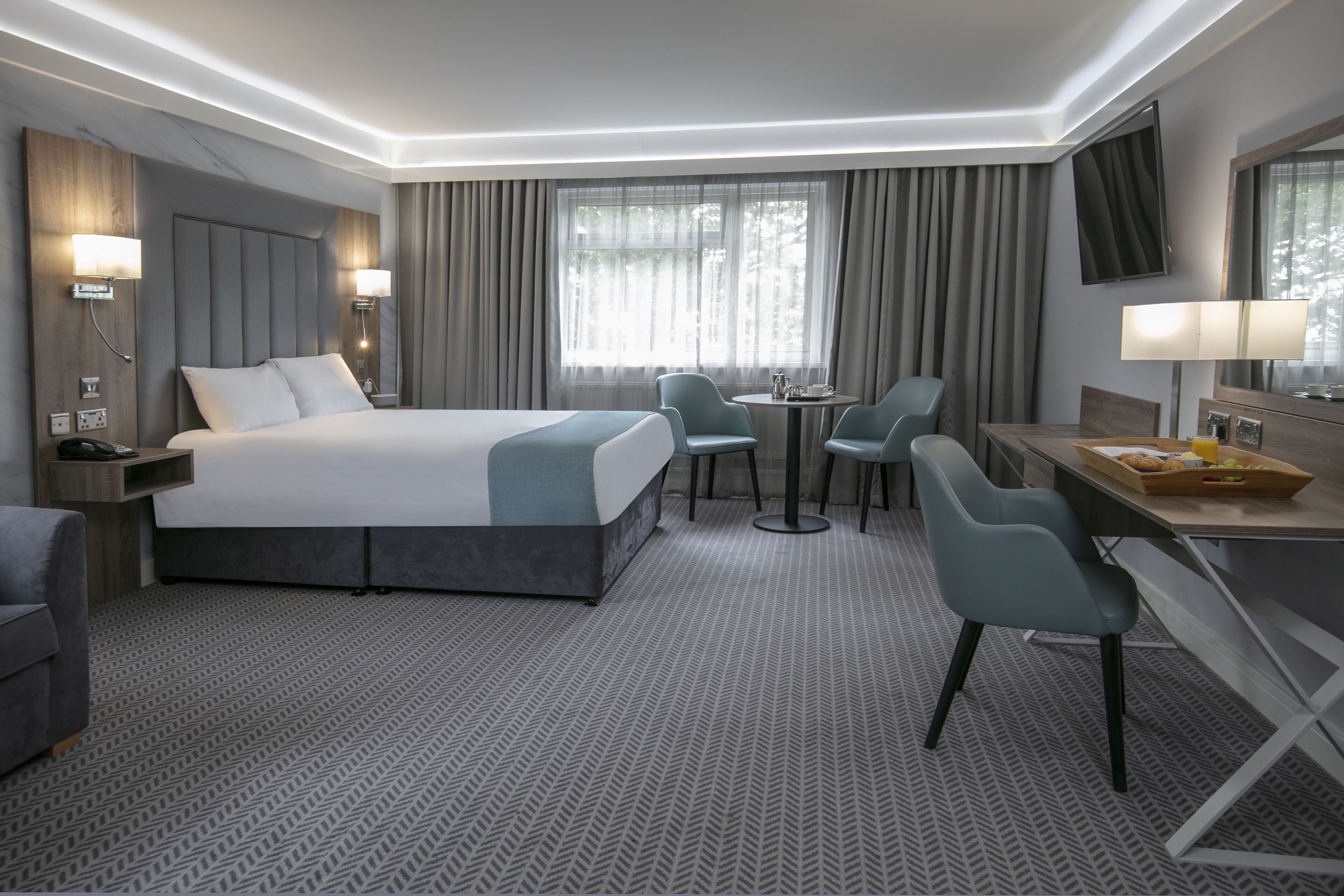 A modern hotel room with a large bed, two chairs, a small table, and a desk with a tray of breakfast items.