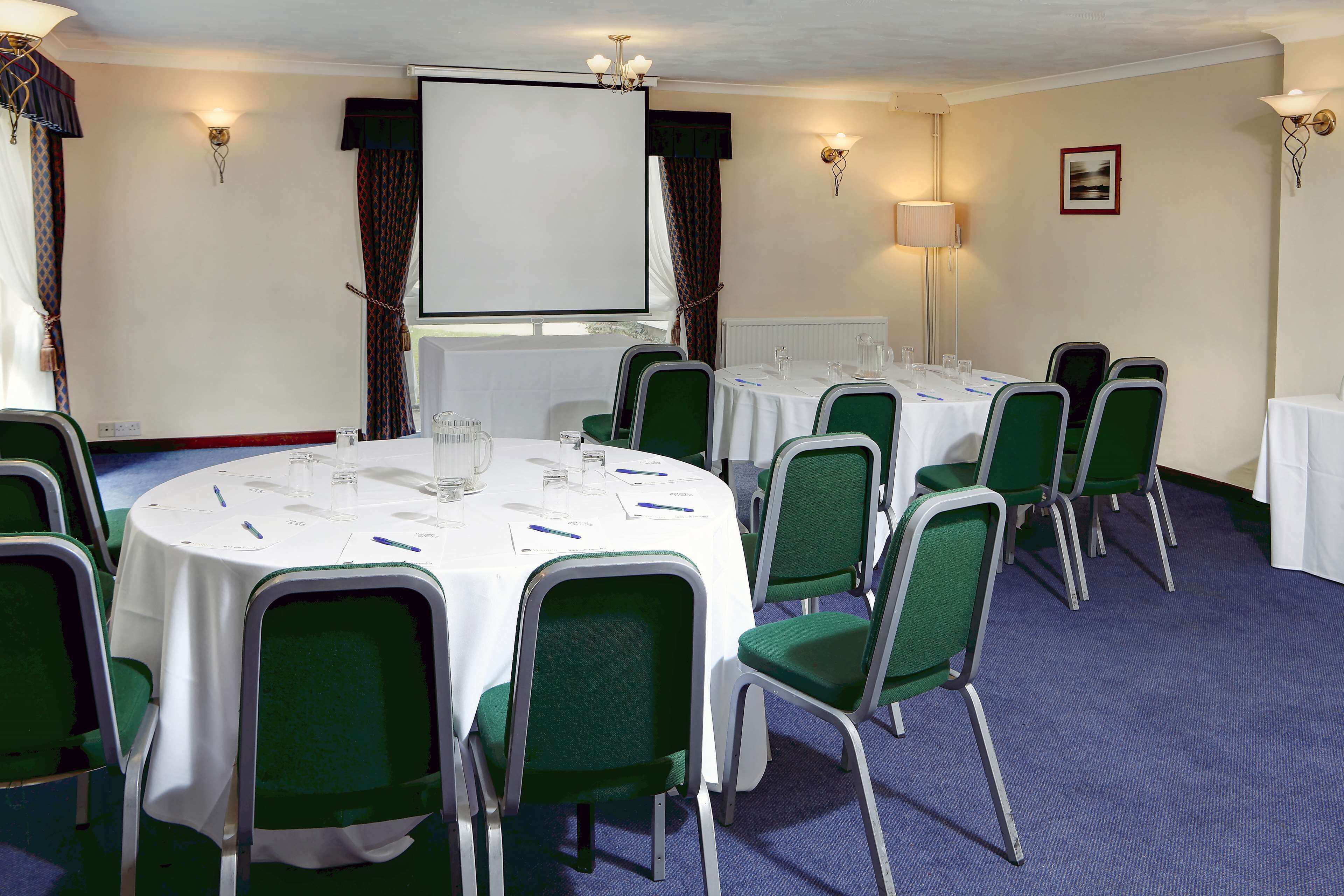 A conference room with round tables, green chairs, white tablecloths, a projector screen, and wall-mounted lamps.