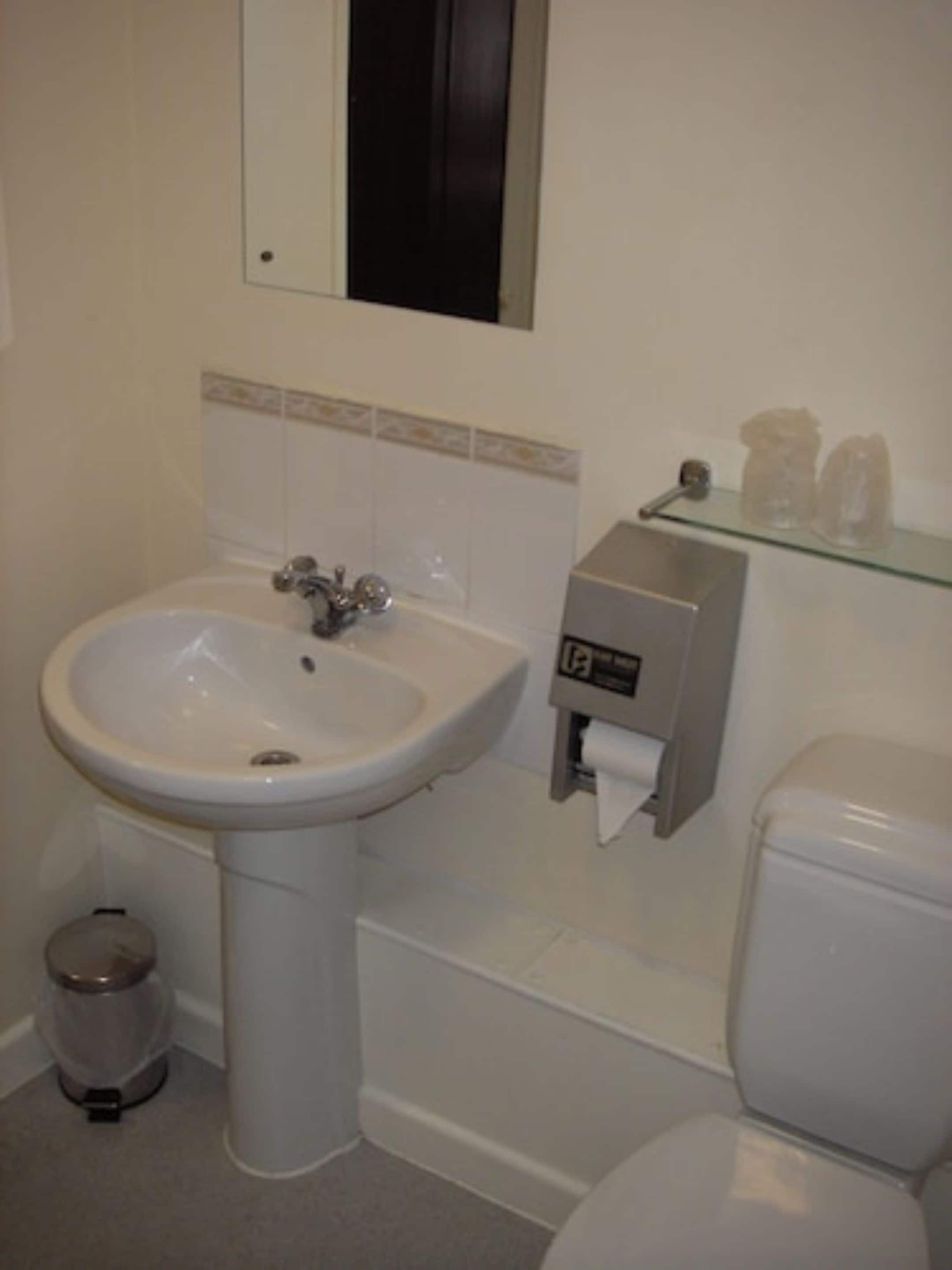 A bathroom with a white sink, toilet, and bidet. There is a trash can and a glass shelf with toiletries.