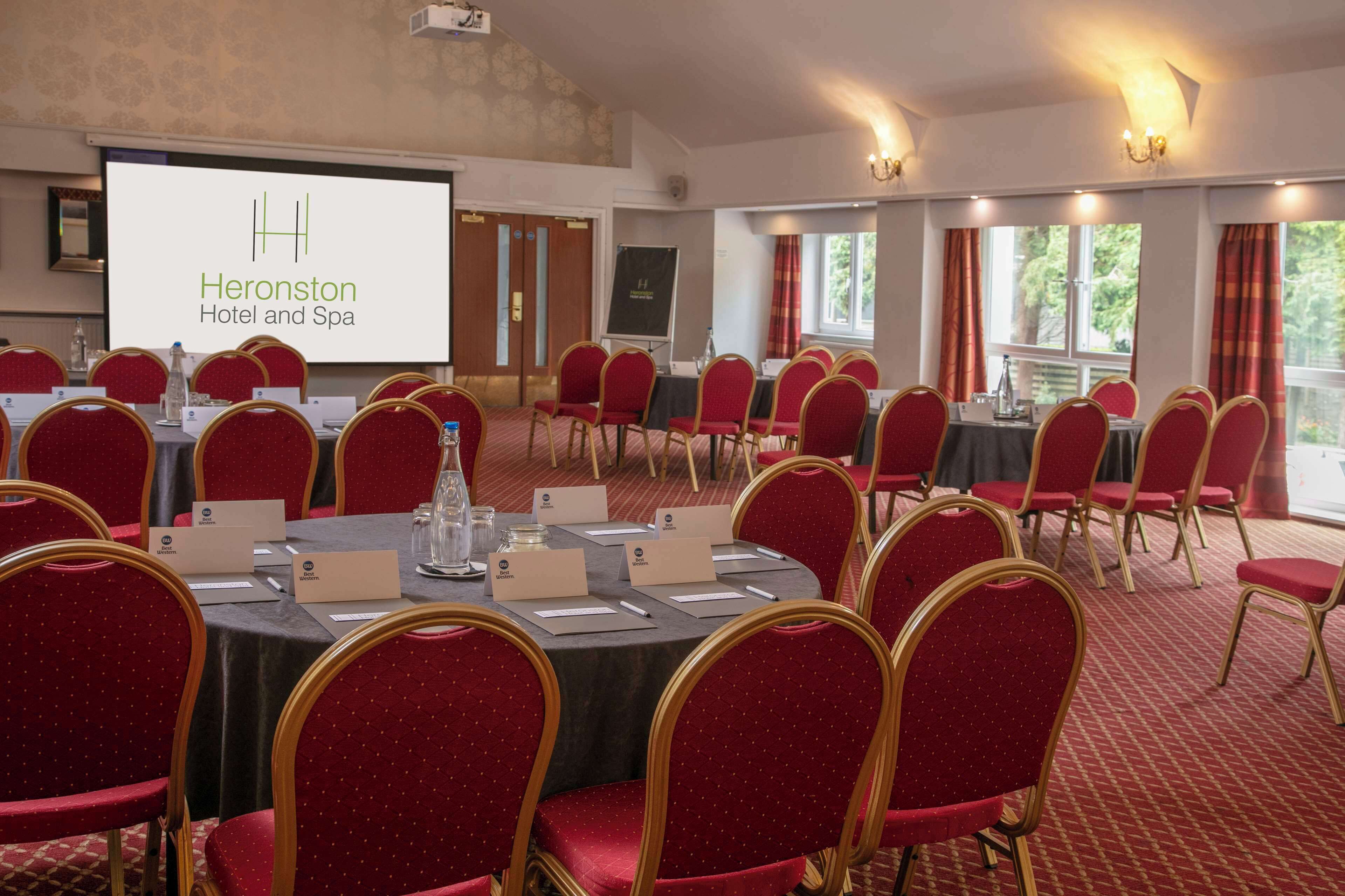 Un salón de conferencias con mesas redondas, sillas rojas, tarjetas de identificación y una pantalla proyectando "Heronston Hotel and Spa".