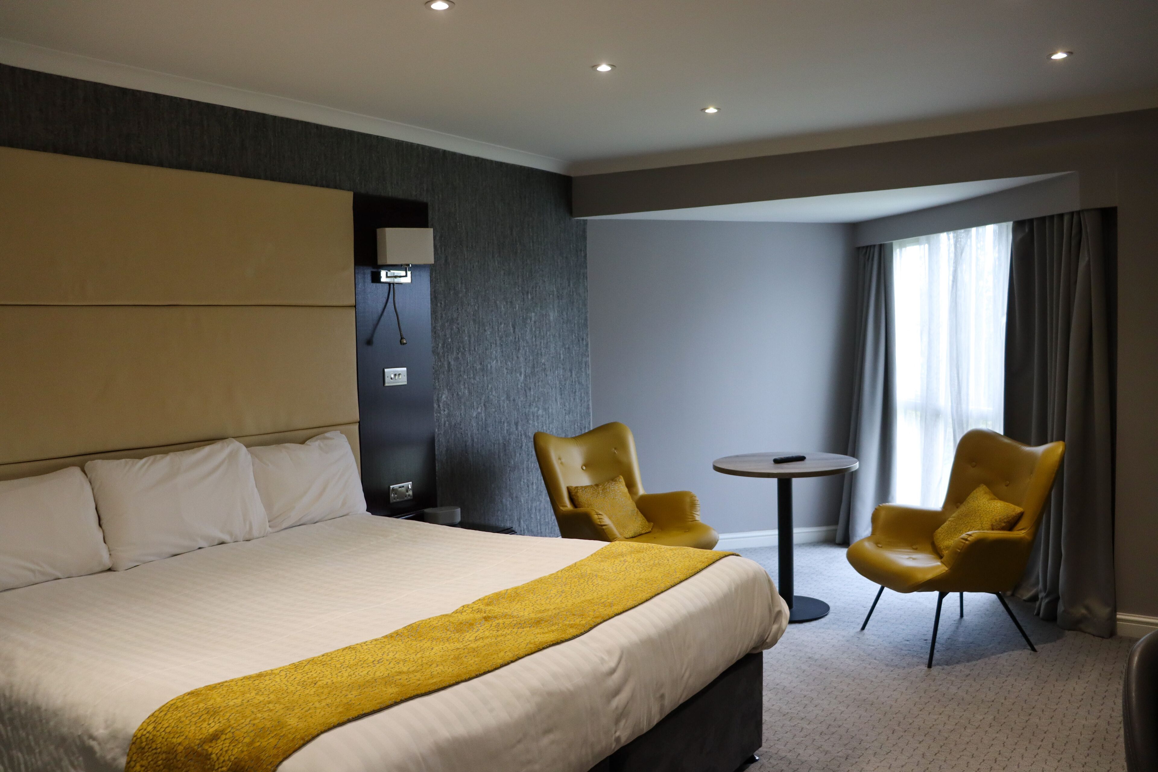 Quarto de hotel com uma cama grande, duas poltronas, uma mesinha redonda e vista para o exterior pela janela.