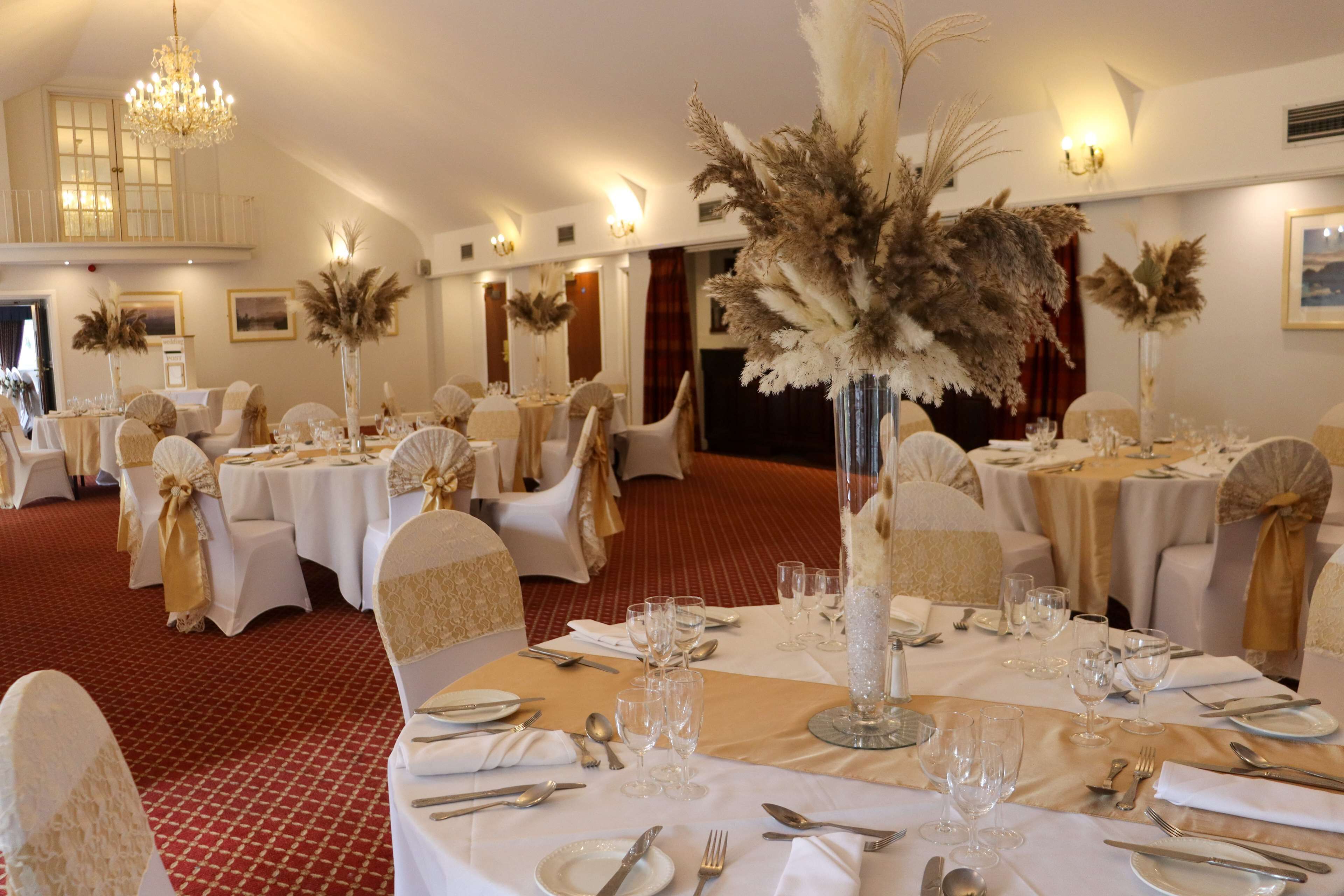 Un salón de banquetes con mesas redondas dispuestas para cenar, sillas blancas con cintas doradas y altos jarrones con flores secas.