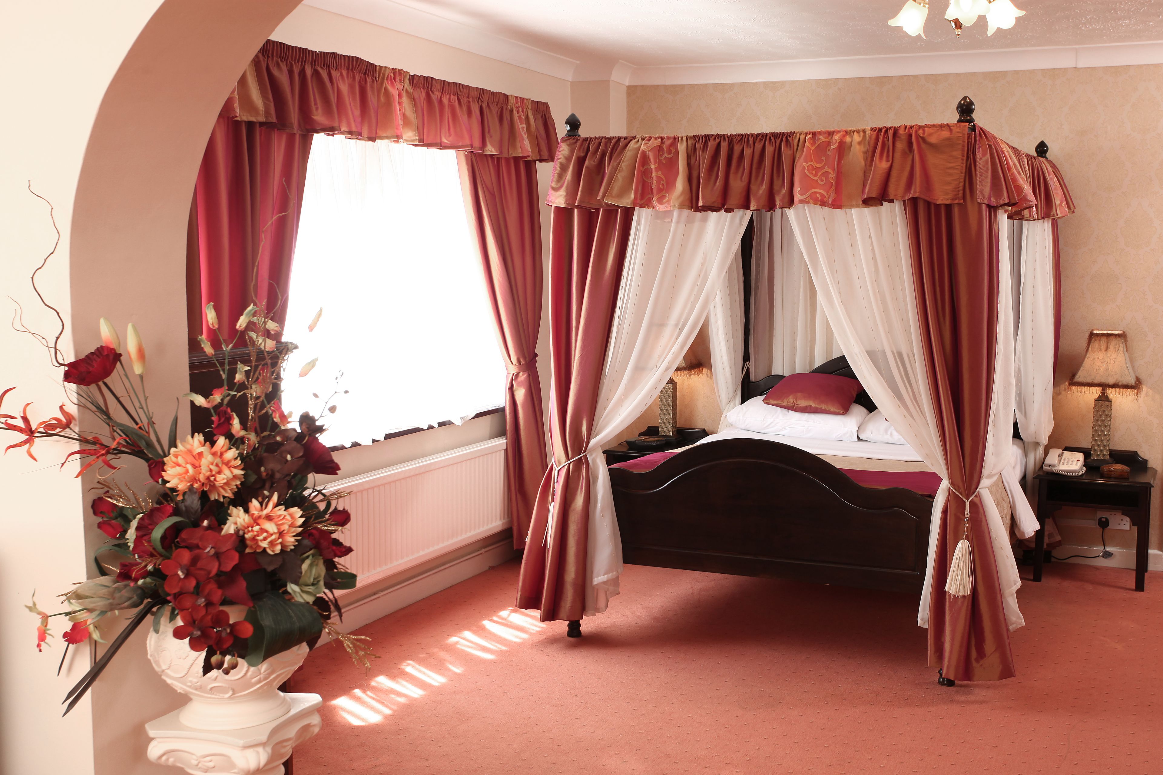 A four-poster canopy bed with a red and white drape, a bedside table with a lamp, and a floral arrangement on a white pedestal.