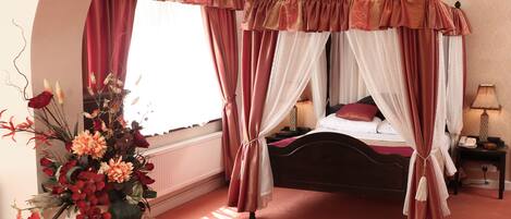 A four-poster canopy bed with a red and white drape, a bedside table with a lamp, and a floral arrangement on a white pedestal.