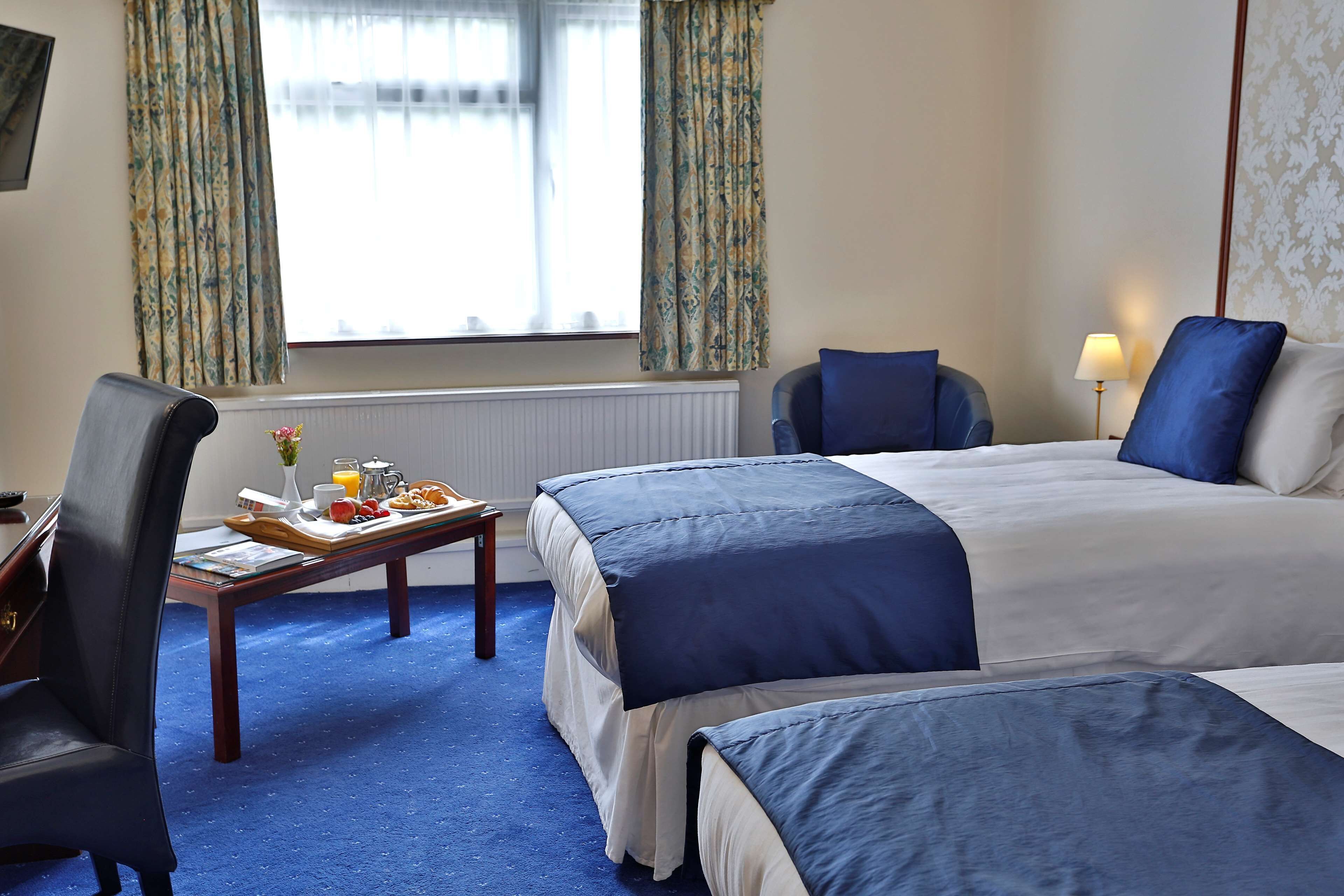 A hotel room with two beds, a desk with a chair, a table with breakfast items, and a window with curtains.