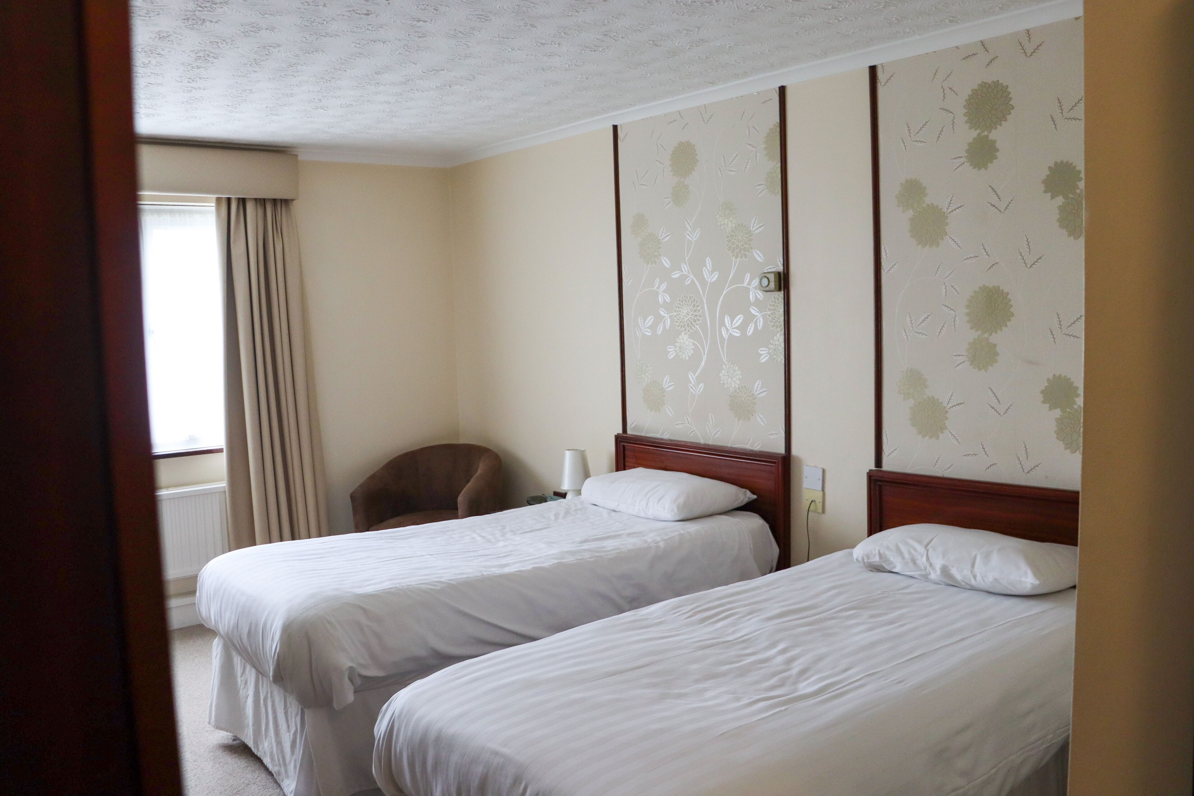 A hotel room with two single beds, a wooden headboard, a chair, a nightstand, and a window with curtains.