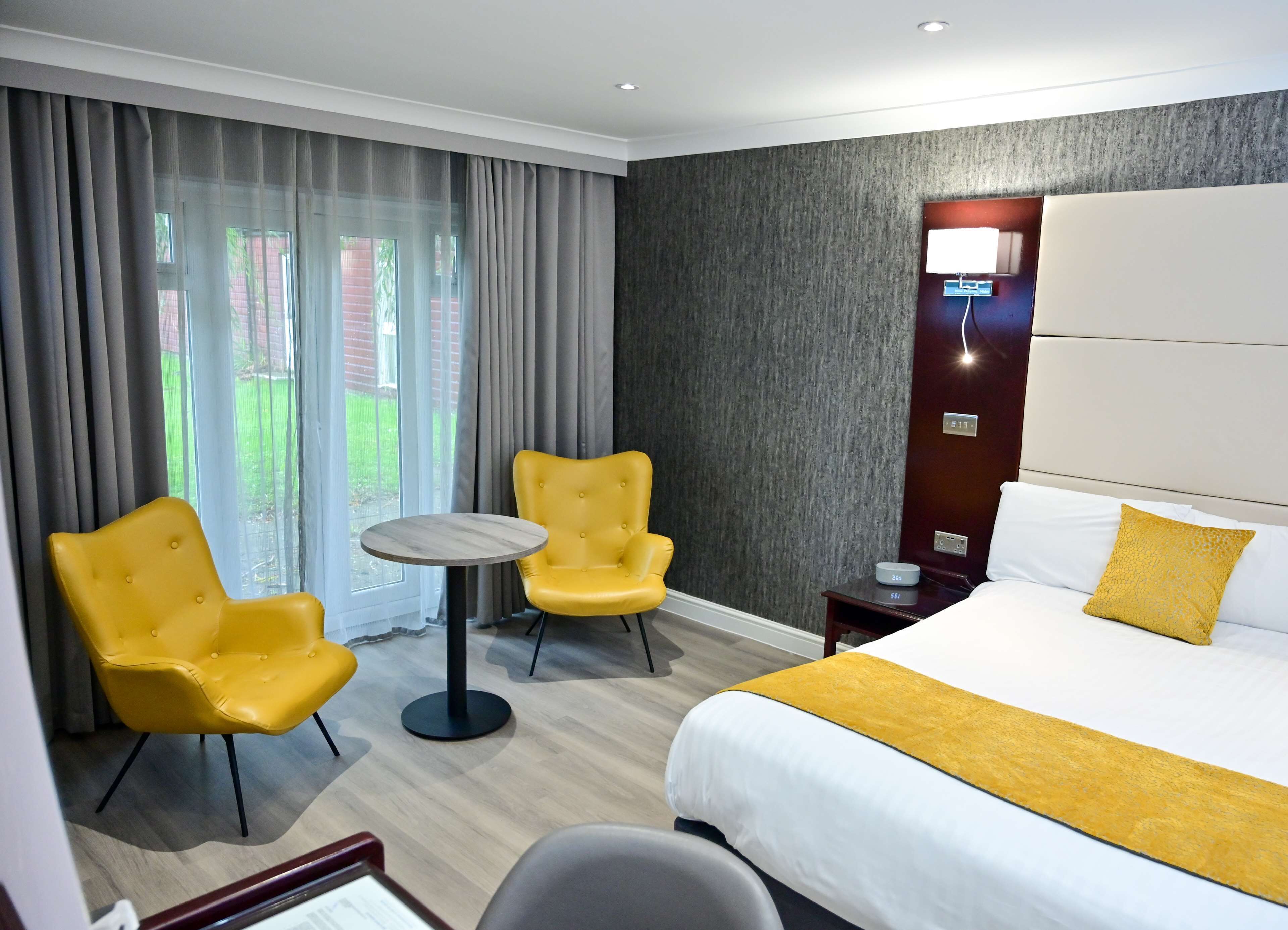 A hotel room with a bed, two yellow armchairs, a small round table, a nightstand, and a view of a green garden through large windows.