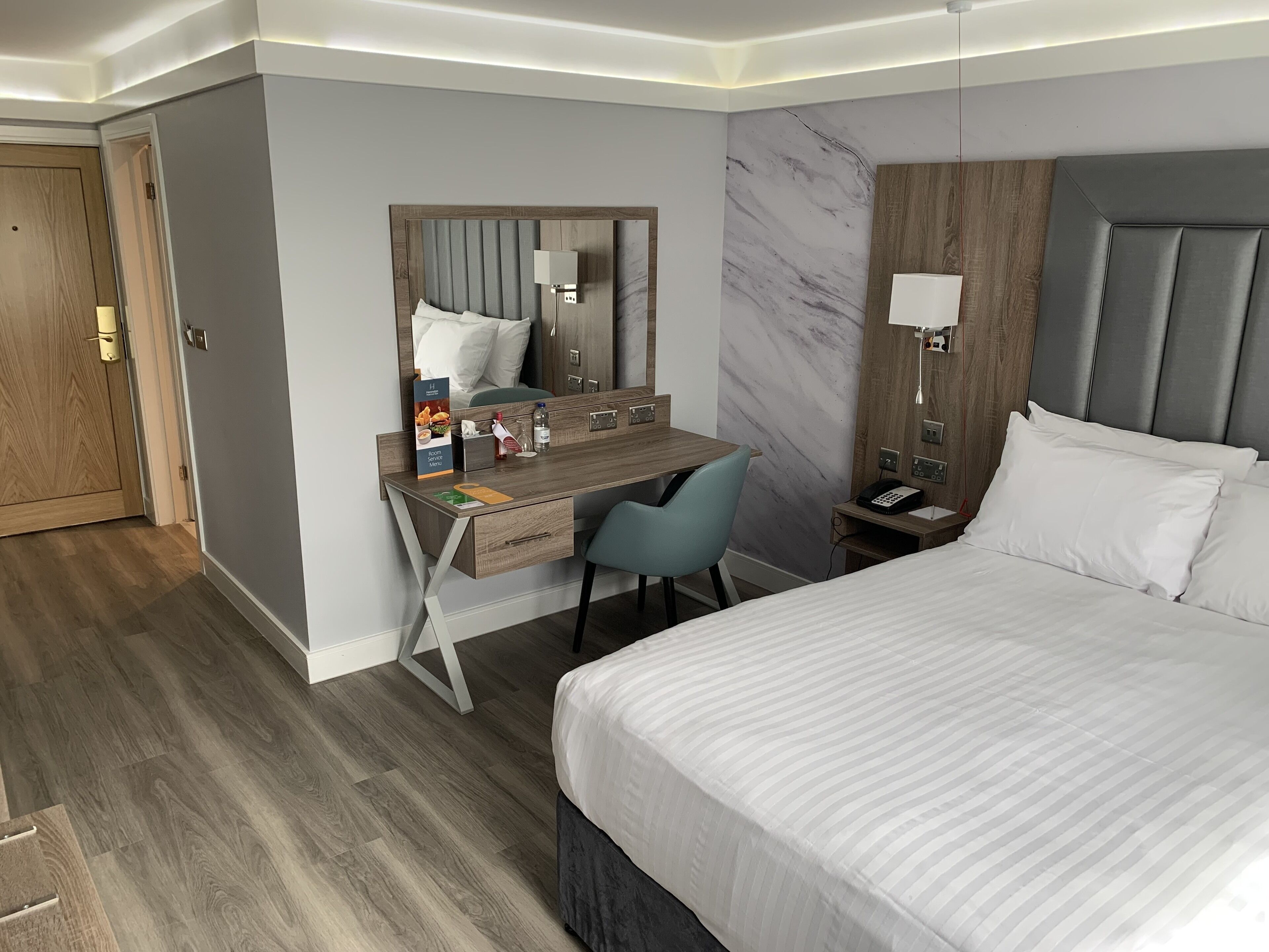 A hotel room with a bed, a desk with a chair, a nightstand, a mirror, and a marble wall.