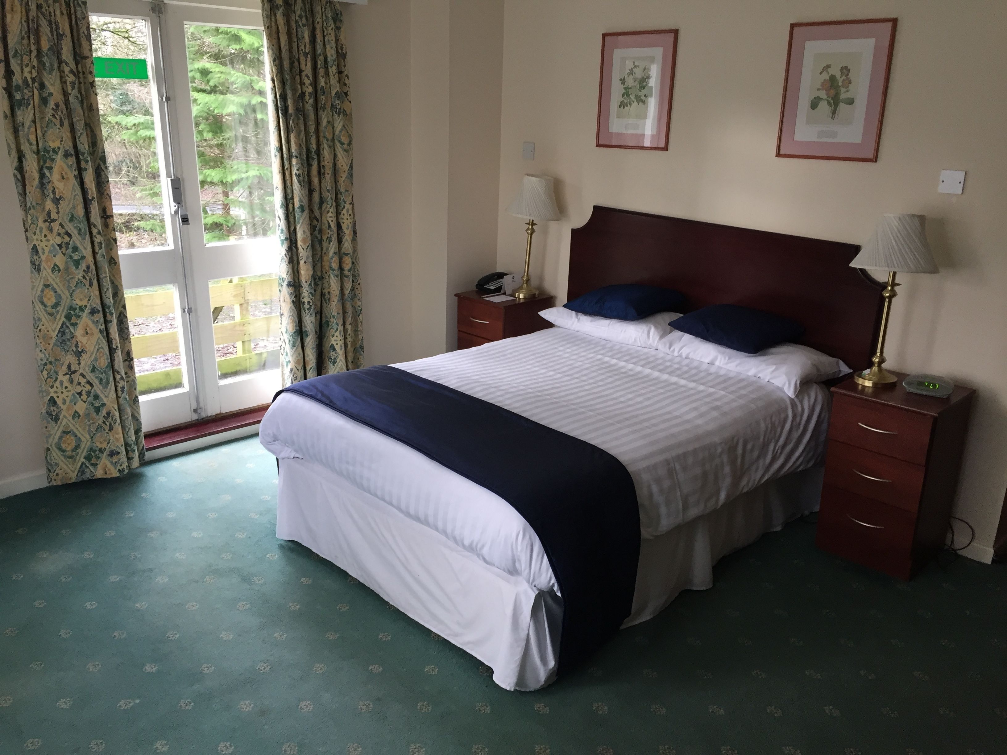 A neatly made bed with a dark blue bedspread, two pillows, and a wooden headboard. There are two bedside tables with lamps and a small clock. The room has a green carpet and a window with curtains.