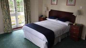 A neatly made bed with a dark blue bedspread, two pillows, and a wooden headboard. There are two bedside tables with lamps and a small clock. The room has a green carpet and a window with curtains.