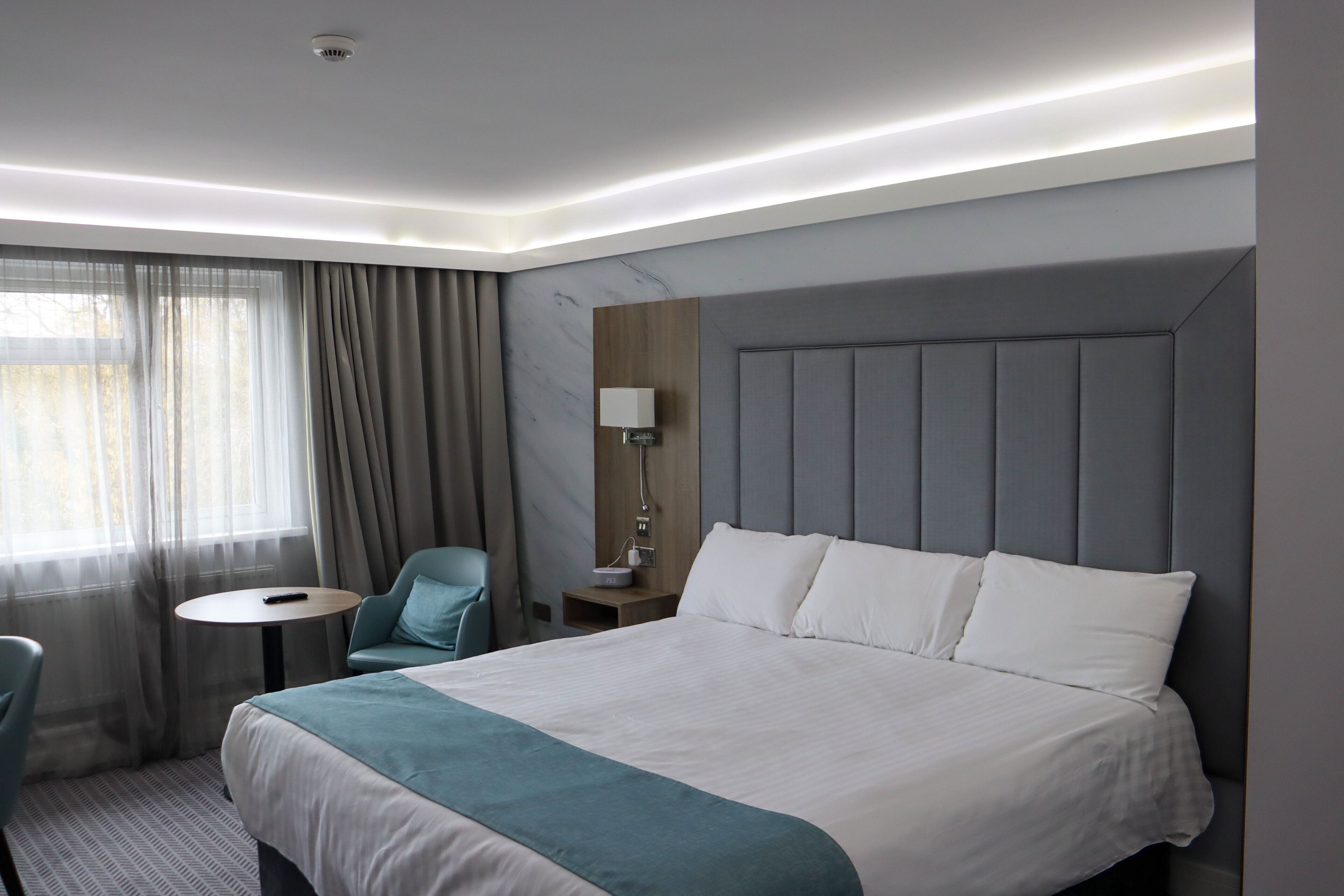 A modern hotel room with a large bed, a small round table, a chair, and a desk with a lamp.