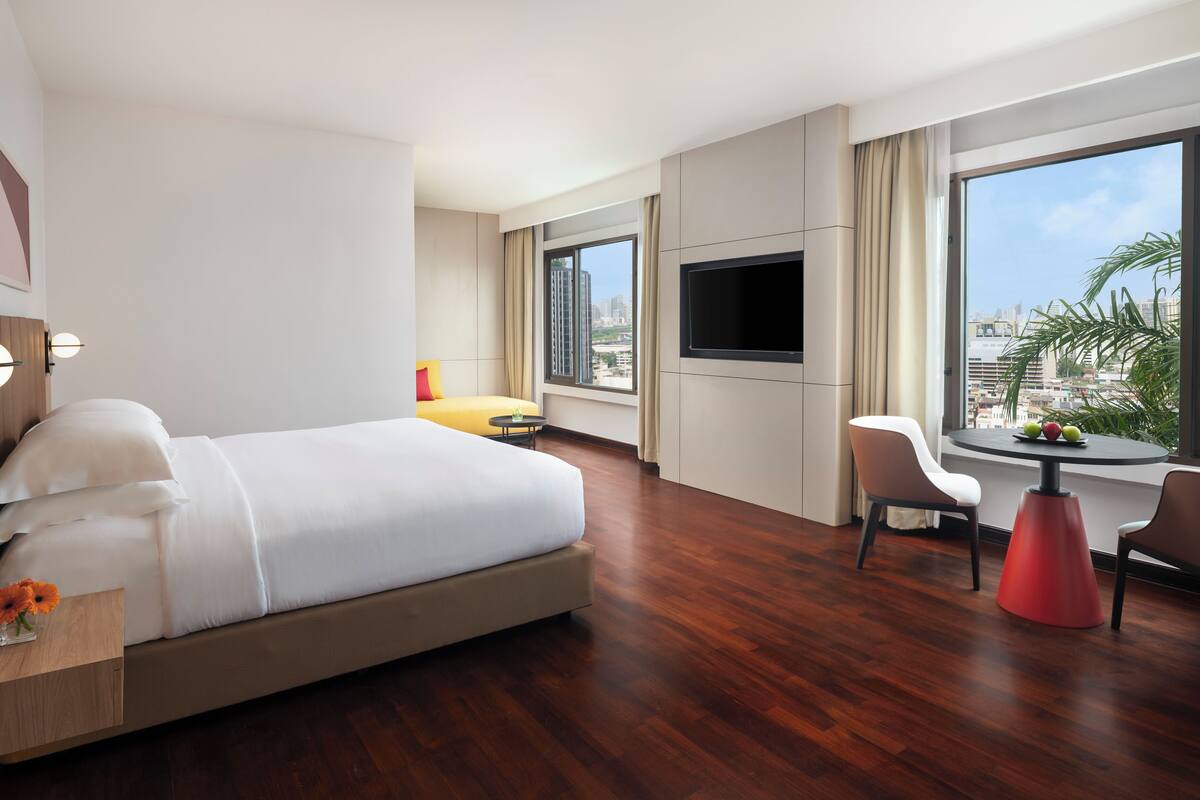 A modern hotel room with a large bed, a TV mounted on the wall, a dining table with chairs, and a view of the city through large windows.